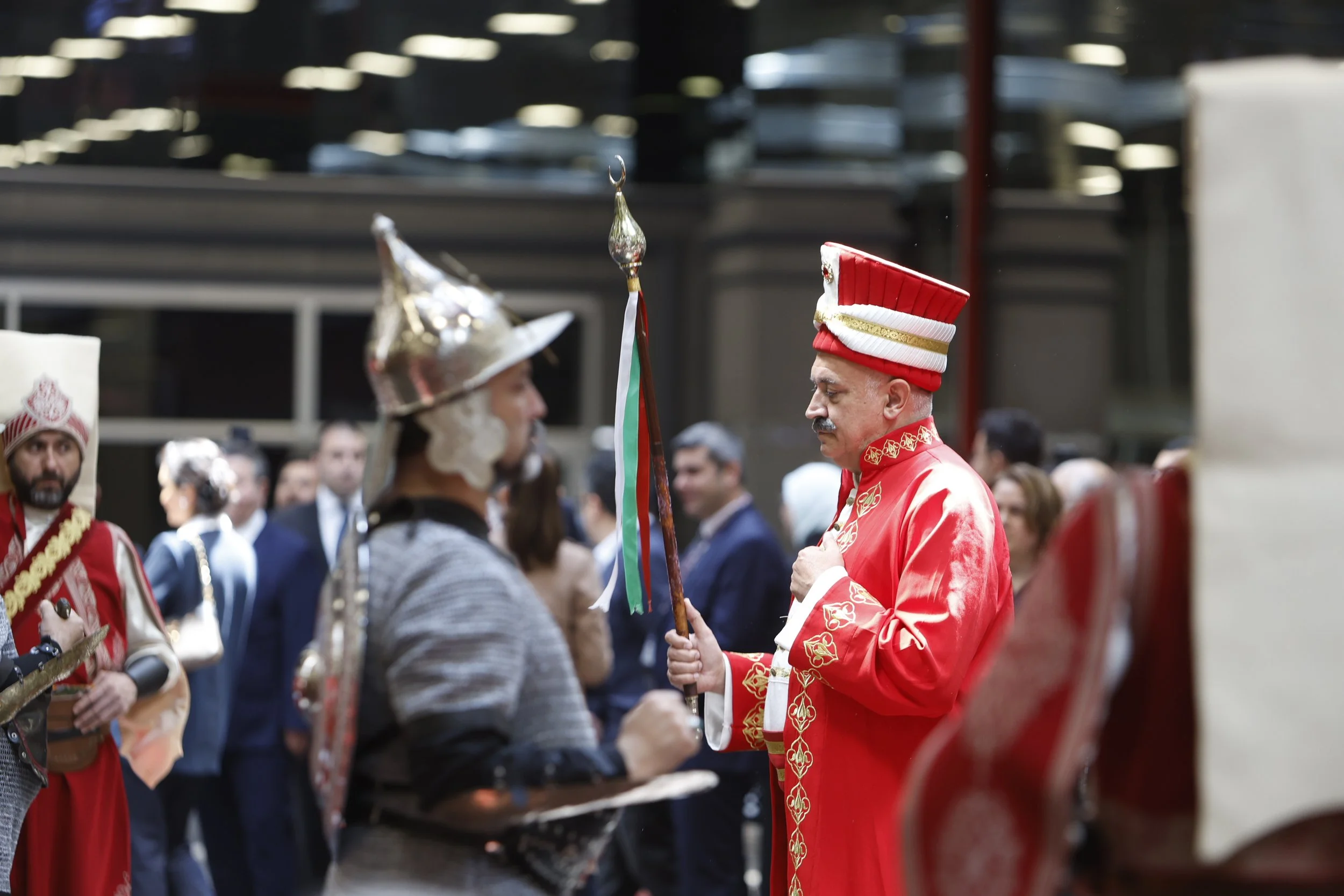 A man dressed in traditional red ceremonial attire with a tall hat, holding a flag, standing in front of a man wearing armor and a metallic hat, amidst a crowd in formal and ceremonial clothing.