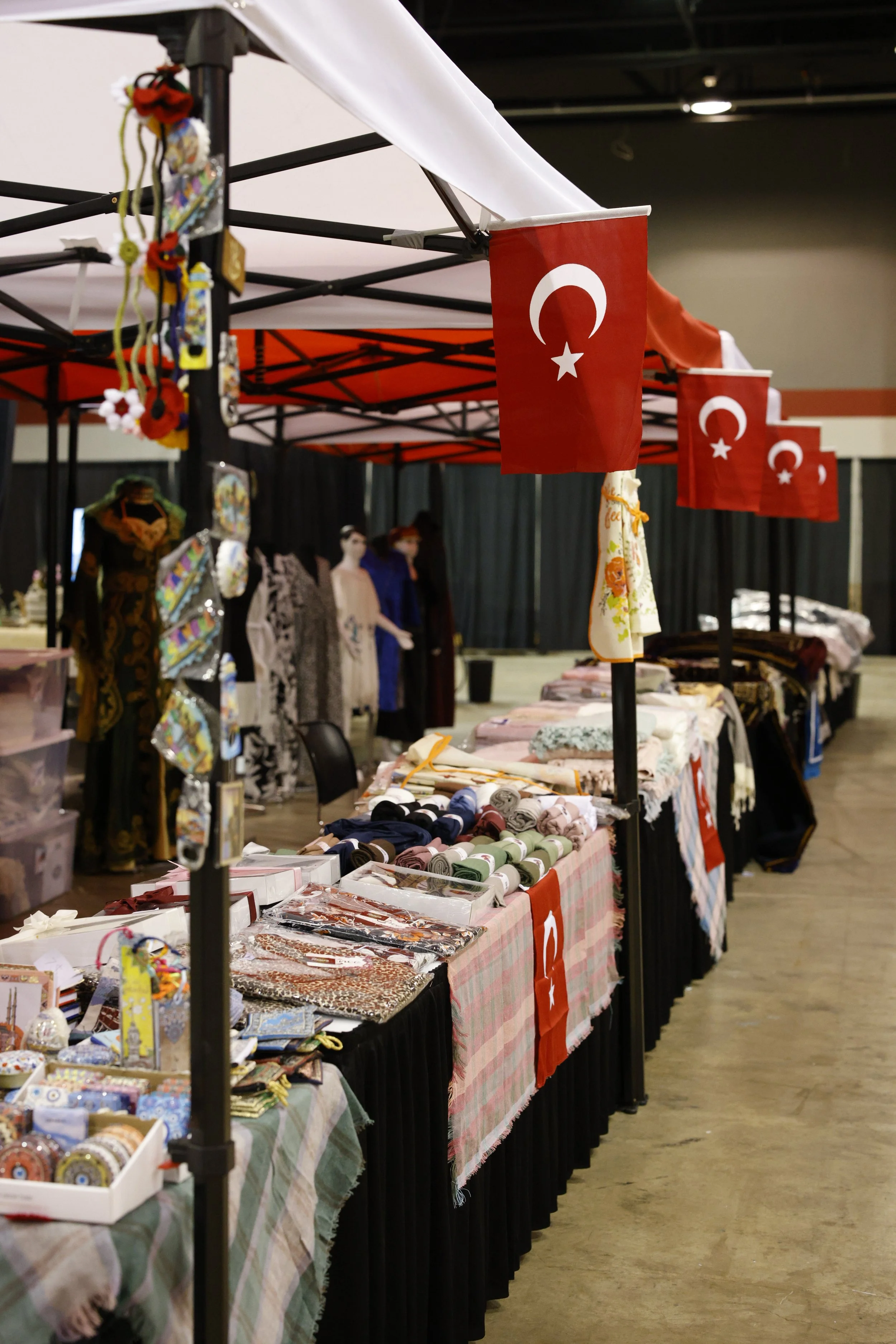 Market stall decorated with Turkish flags, displaying various textiles, clothing, and accessories for sale. Mannequins and hanging garments are visible in the background.