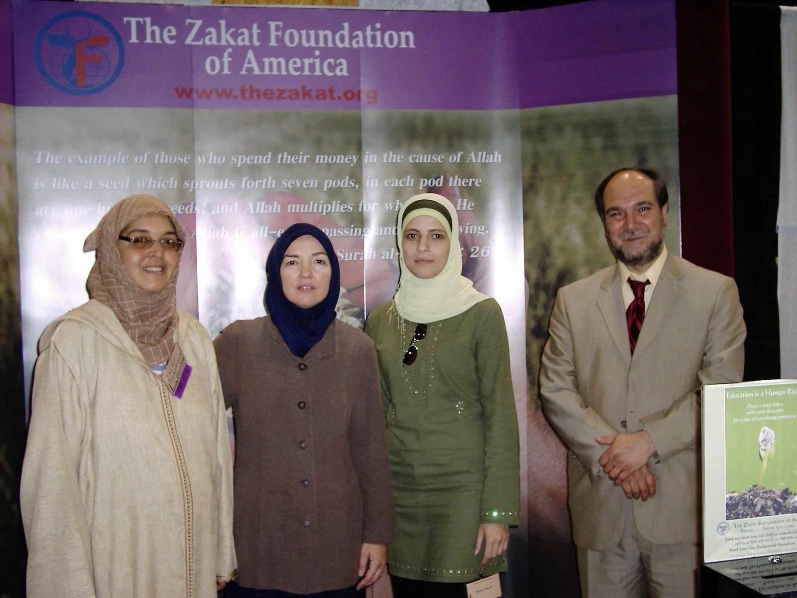 Group of four people standing in front of a backdrop for The Zakat Foundation of America, with a quote from the Quran visible behind them.