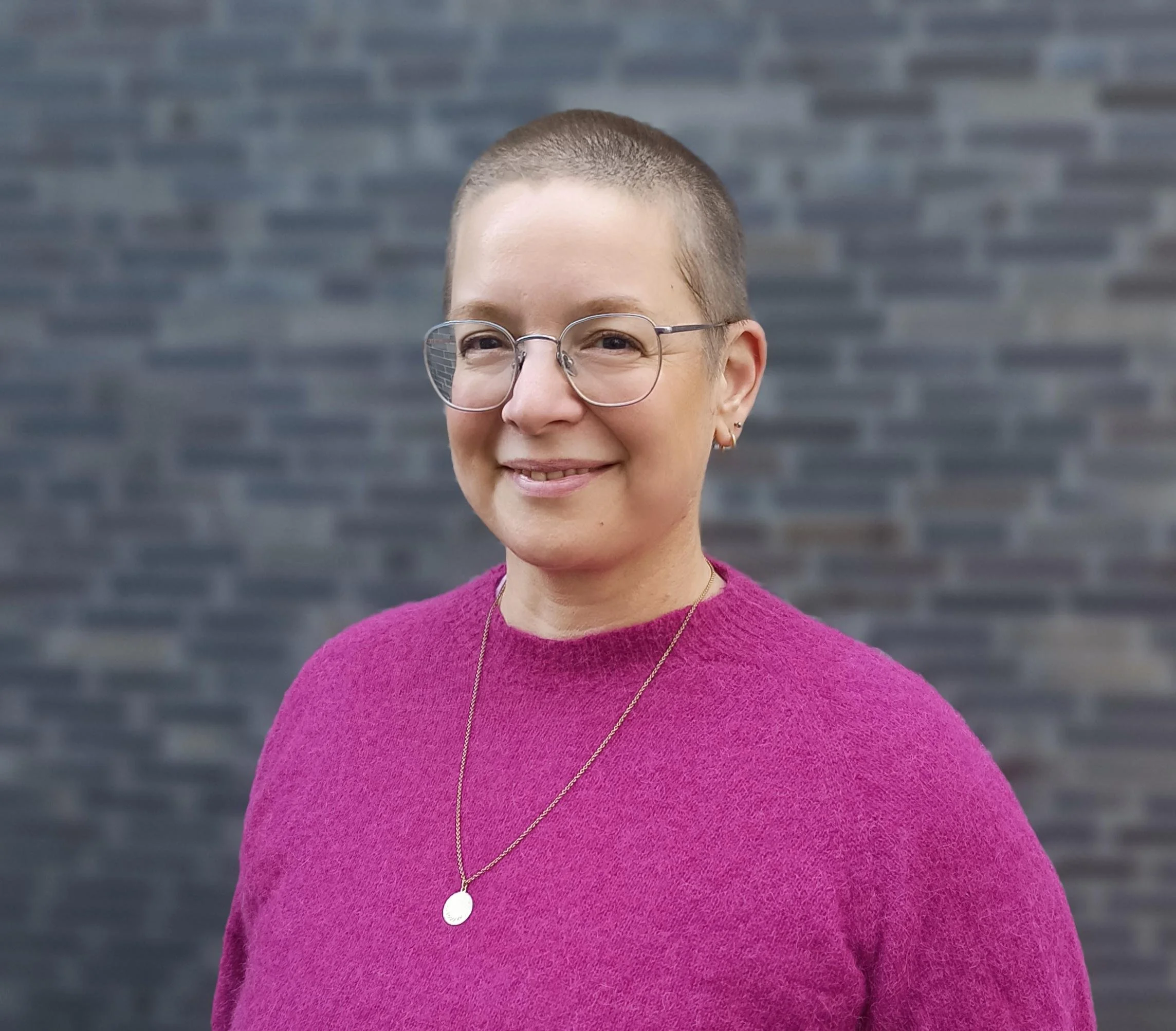 A woman with short hair, glasses, wearing a pink sweater and a gold necklace, standing in front of a dark brick wall.
