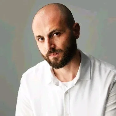 A person with a bald head and beard wearing a white shirt, looking at the camera.