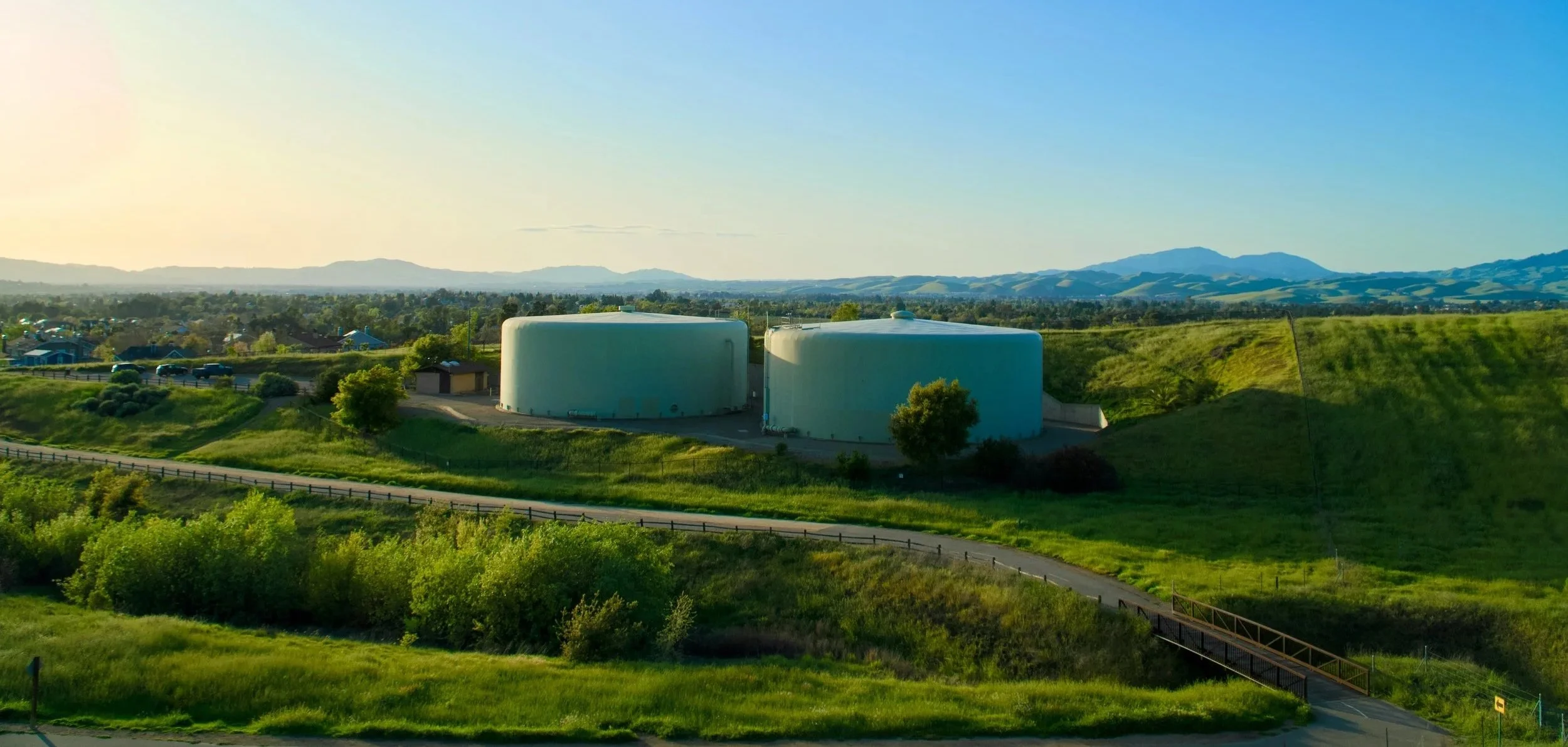 Due grandi serbatoi di liquidi blu su un'area verdeggiante con colline in lontananza e cielo chiaro