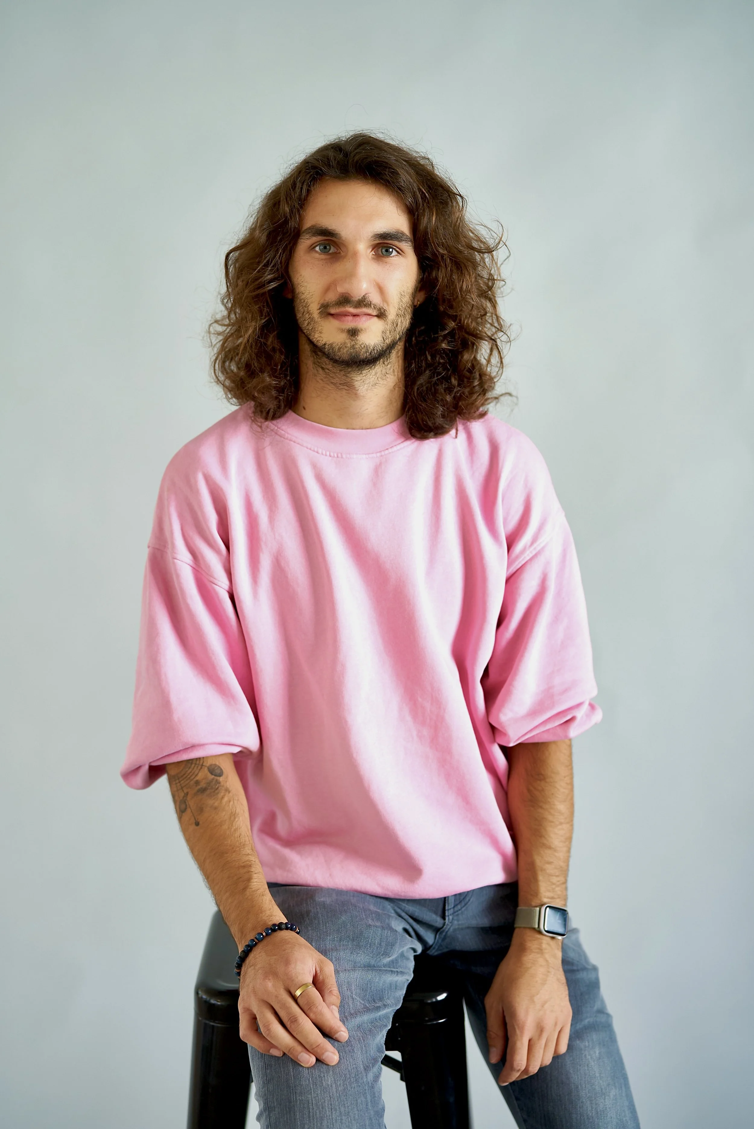 David Arun Sridharan, a man with long curly hair and a beard, wearing a pink t-shirt and gray jeans, seated on a black stool against a plain light-colored background.