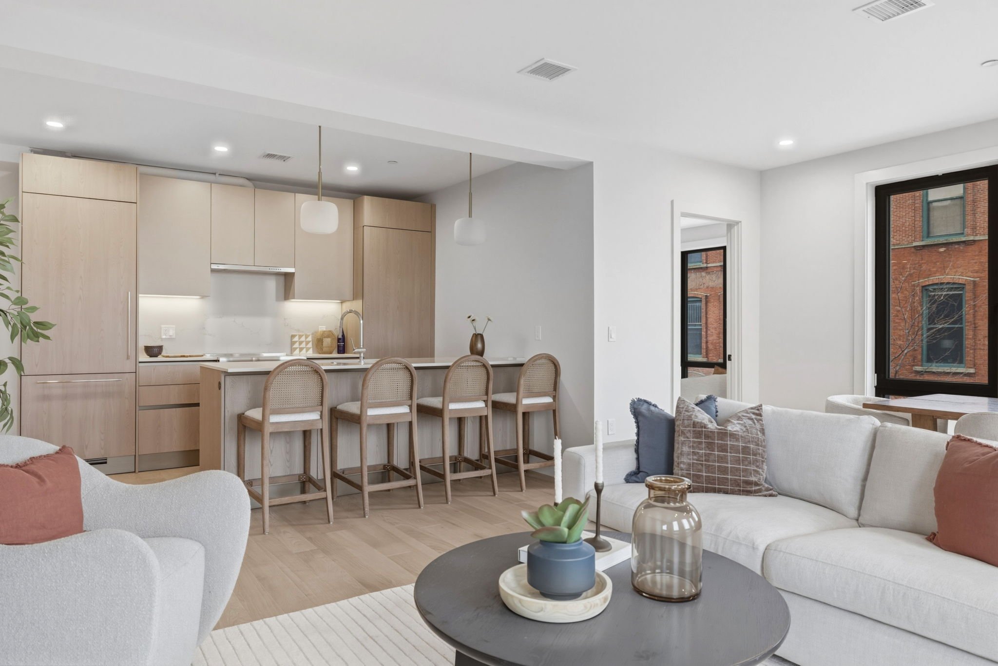 Open-concept living room and kitchen with white walls, large windows, and modern furniture including a white sofa, beige chairs at the kitchen island, and decorative items on the coffee table.