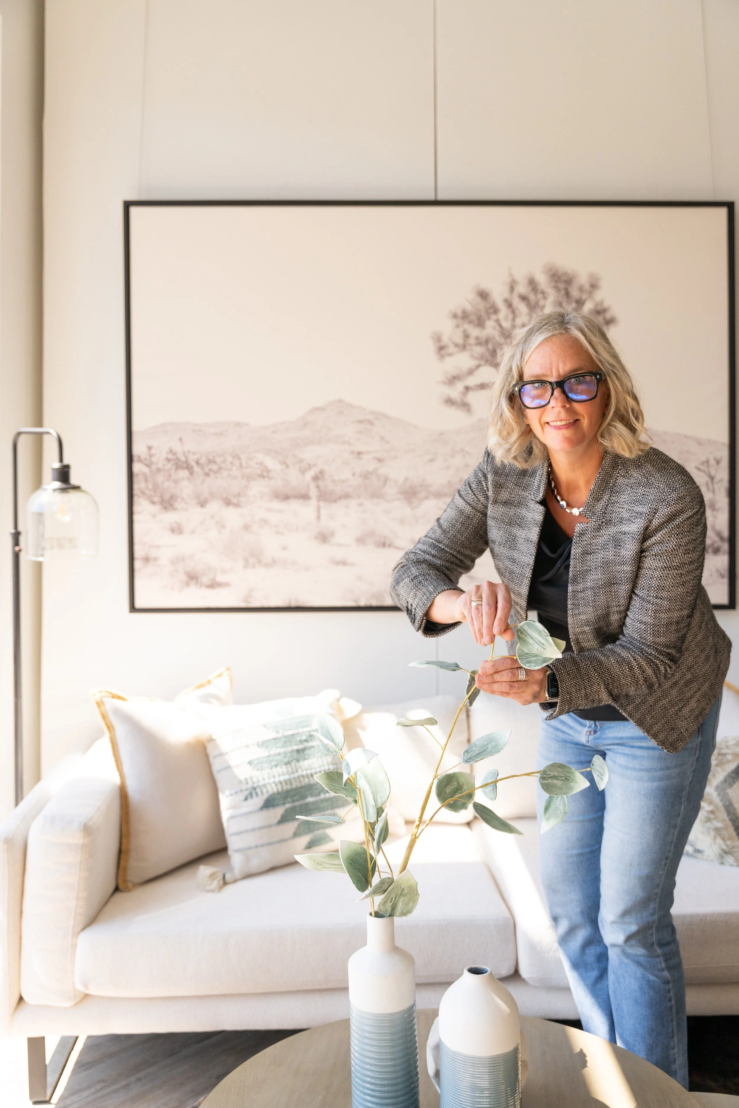 A woman with glasses, curly gray hair, wearing a gray blazer and jeans, standing in a living room arranging a green plant in a white vase, with a large black-and-white landscape photograph on the wall behind her.