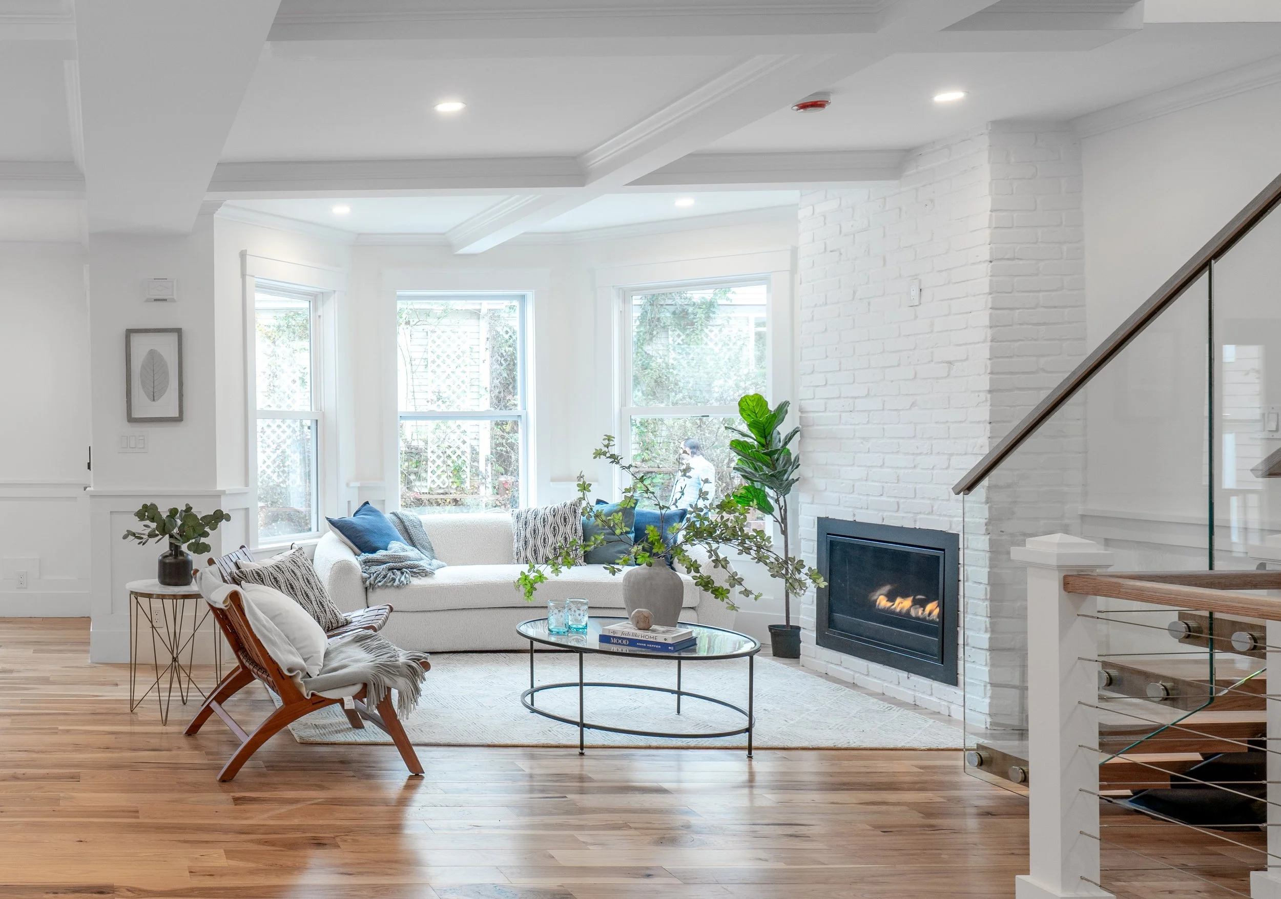 Bright living room with white walls, large windows, a cozy white sofa with blue and gray pillows, a wooden chair with a blanket, a round glass coffee table, a large vase with green plants, a white brick fireplace, and hardwood floors.