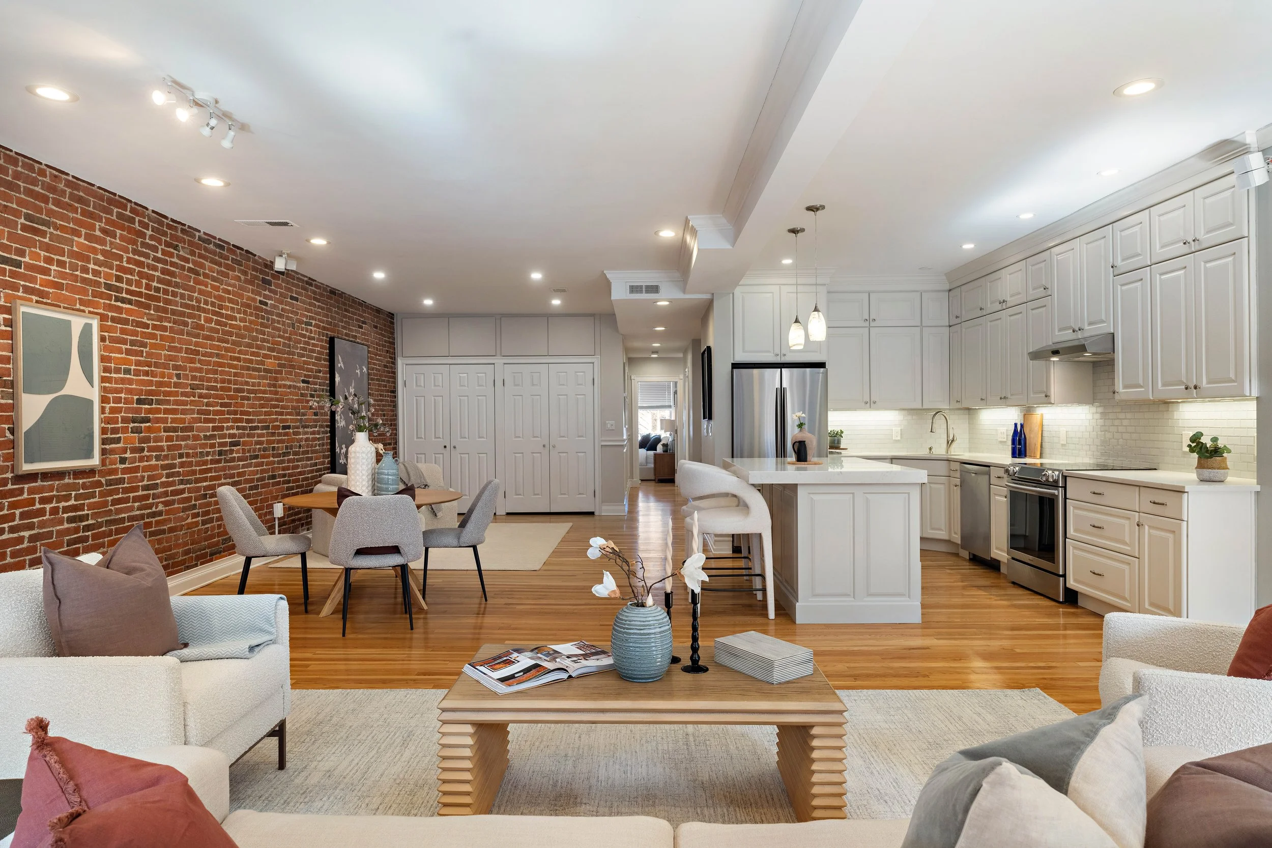 Open-concept living room and kitchen with an exposed brick wall, white cabinetry, stainless steel appliances, a dining table with chairs, and a sitting area with sofas and decorative pillows.