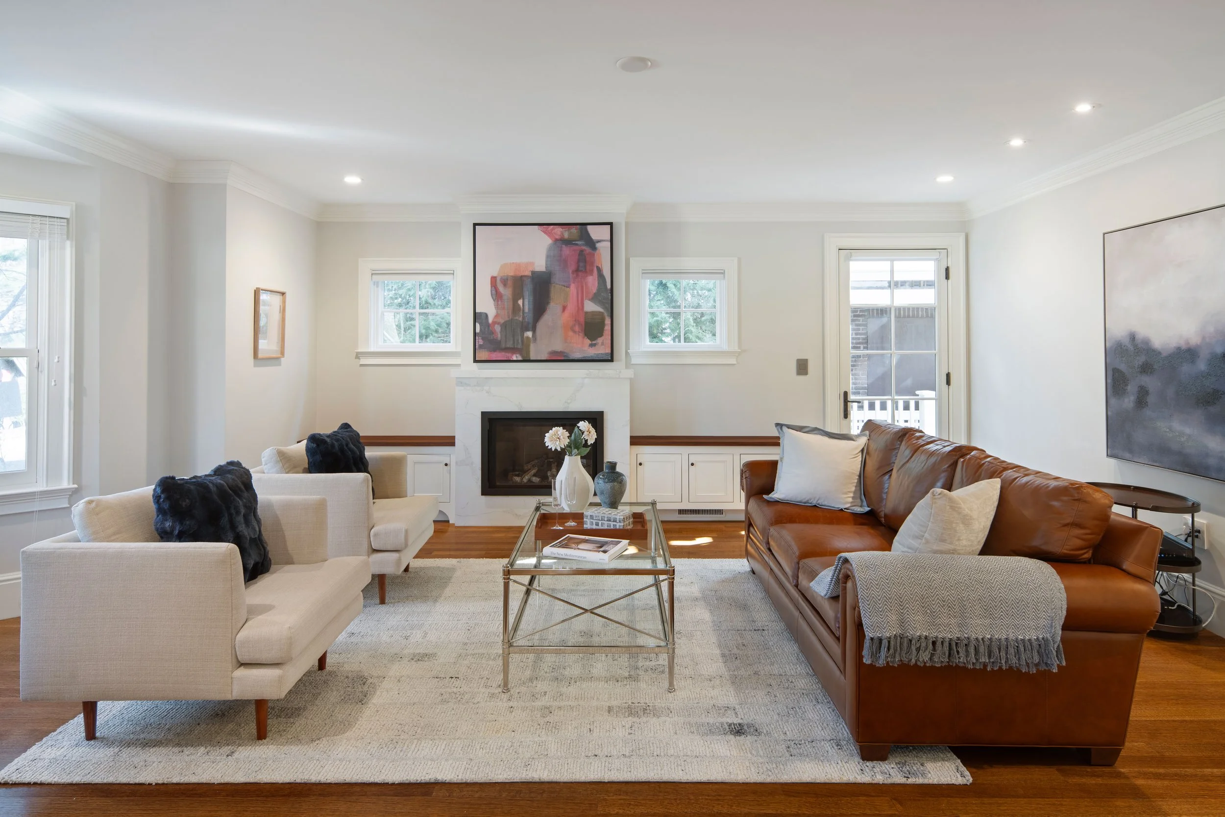 A living room with white walls, hardwood floors, and a white rug. There is a white sofa with black fur pillows, a brown leather sofa, and a glass coffee table with artistic vases. Two small windows and a door leading outside are above a white fireplace with an abstract painting above it. Artworks are on the walls.