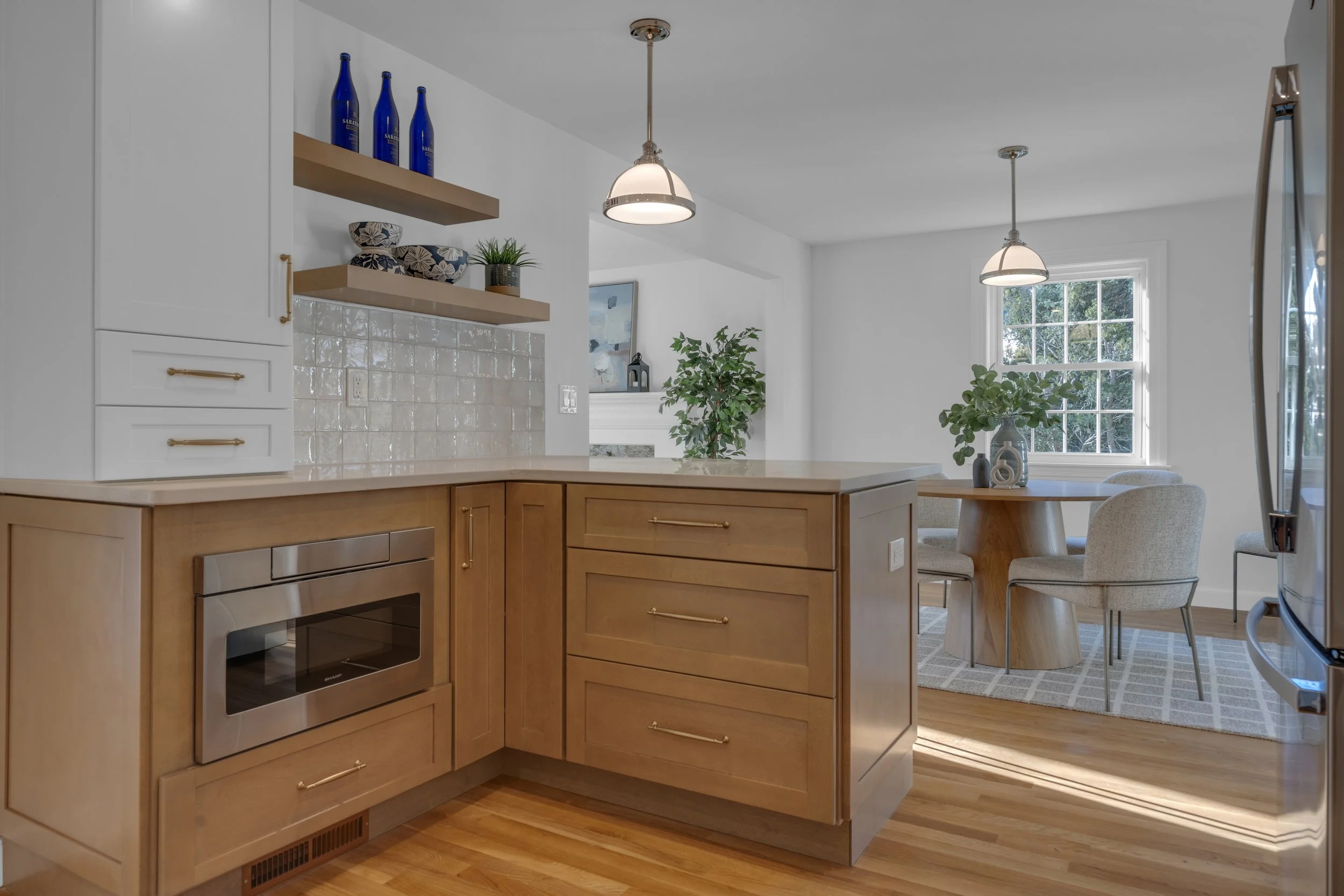 Kitchen with wooden cabinets, white upper cabinets, a microwave, open wooden shelves with blue bottles, plants, a window, and a small dining area with a round table, four chairs, a window with greenery outside, and pendant lights.
