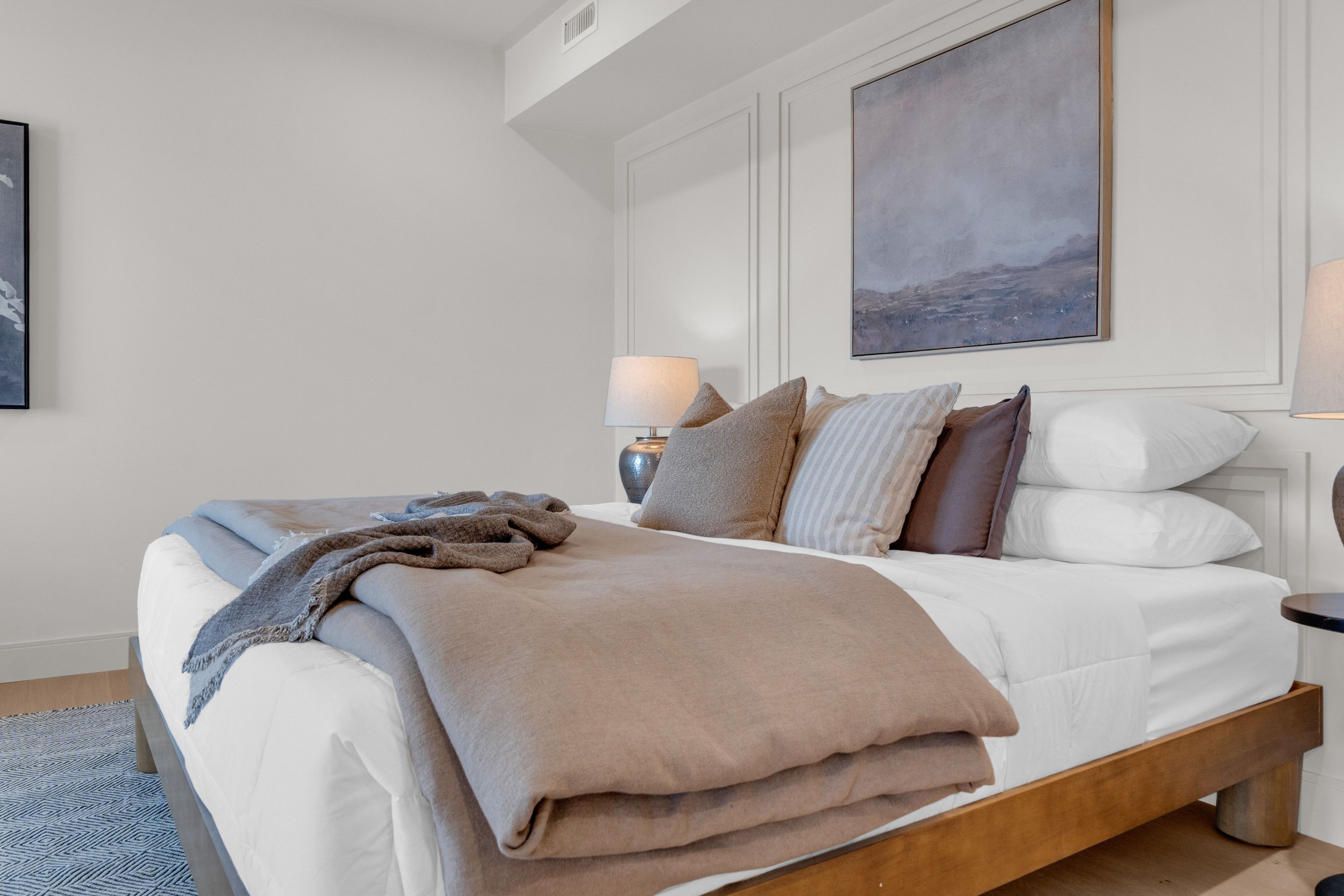 A neatly made bed with a brown blanket and various pillows in shades of beige, gray, and brown, flanked by two lamps on nightstands in a modern bedroom with white walls and framed artwork.
