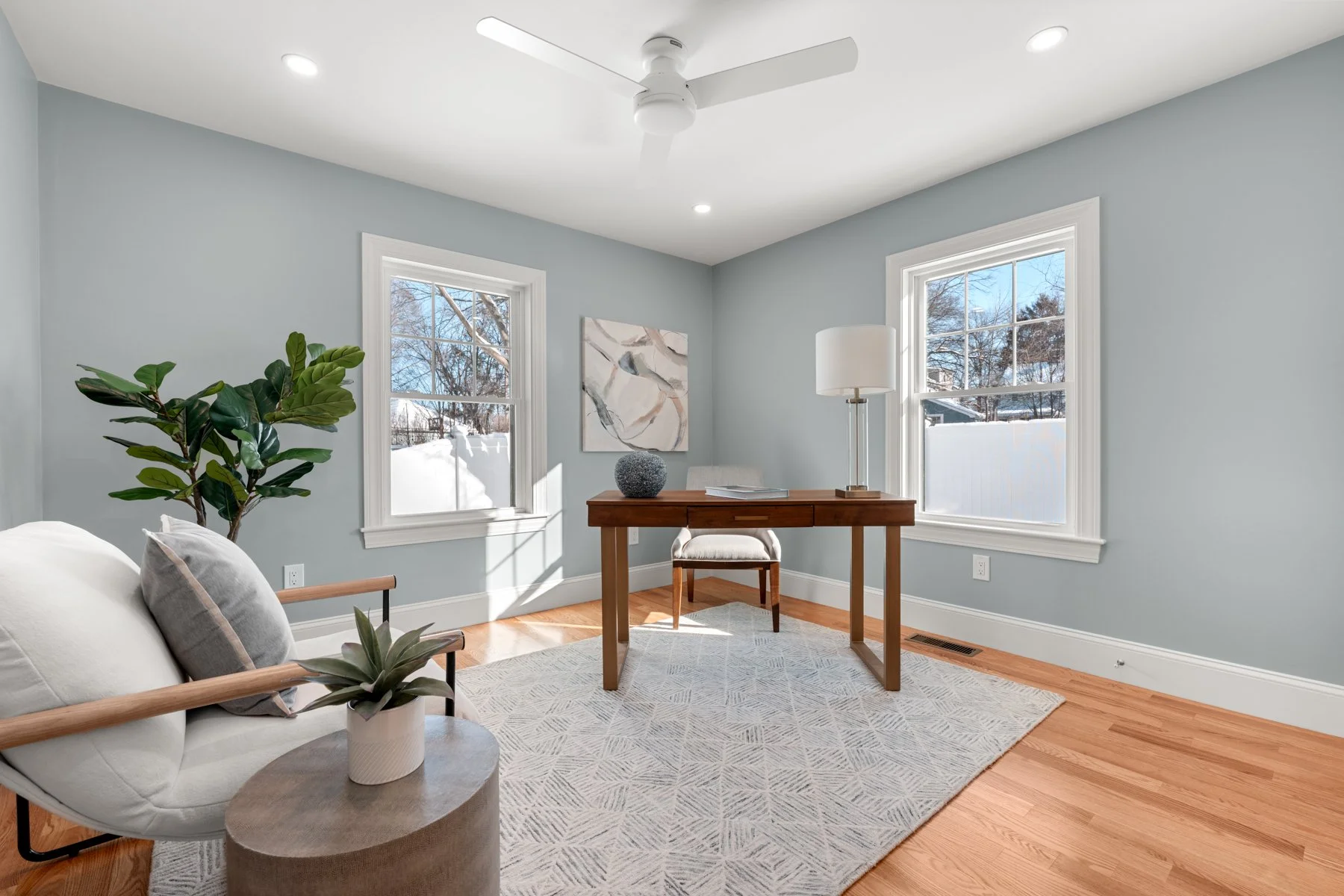 A bright living room with light blue walls, two windows with snow outside, a white ceiling fan, a white armchair with pillows, a plant, a grey rug, and a wooden desk with a lamp and decor.