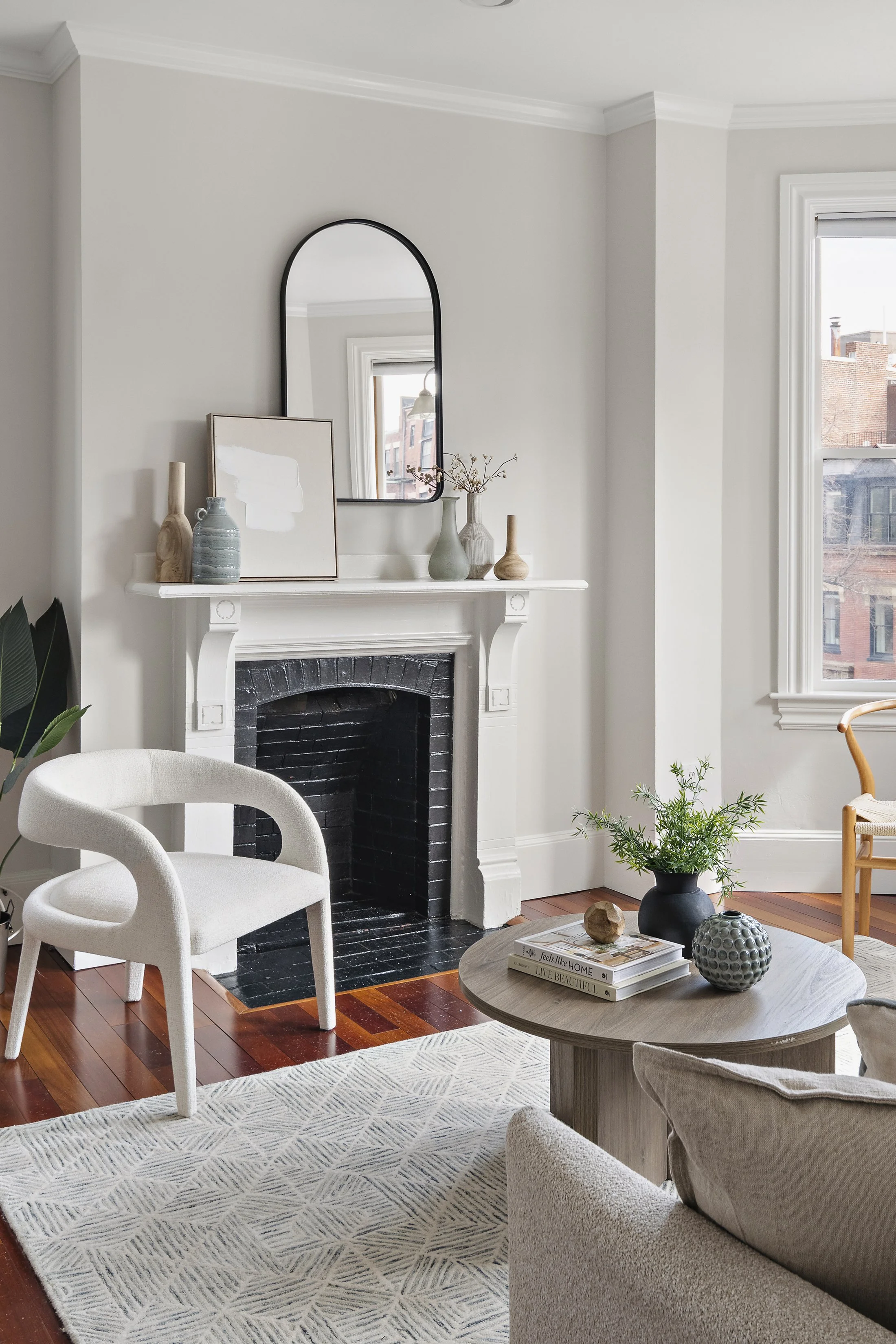 Living room with white walls, a black fireplace, a mirror, artwork, vases, a chair, a coffee table with books and decor, and a large window with natural light.