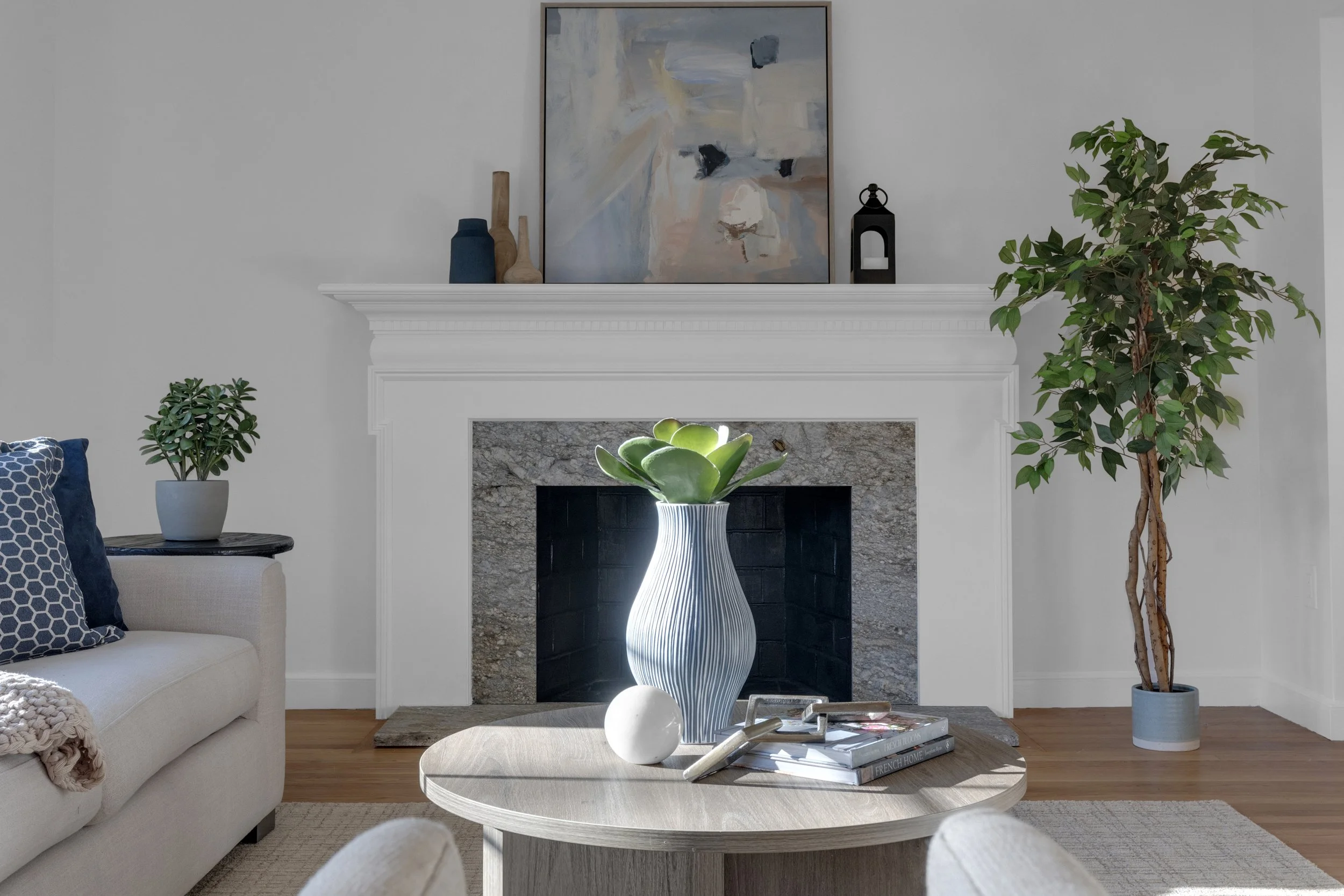Living room with white fireplace, abstract painting, potted plants, a round coffee table with a white vase, books, and decorative objects.