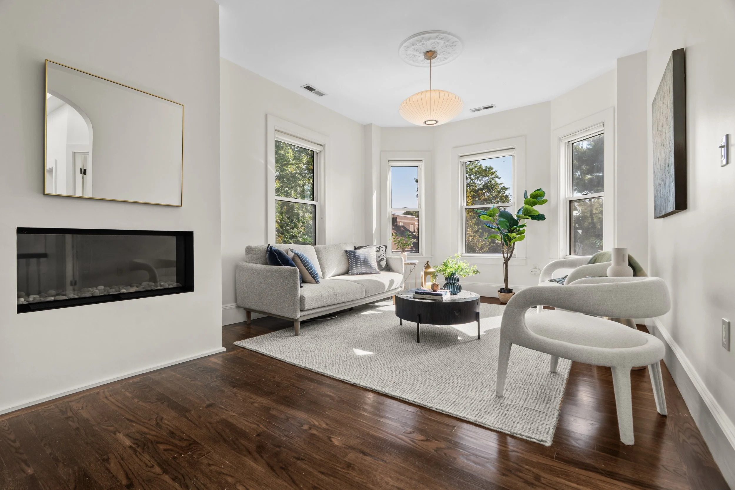 Bright living room with white walls, hardwood floors, a beige sofa with blue pillows, two white modern chairs, a black round coffee table with decorative items, potted plant, and large windows letting in natural light.