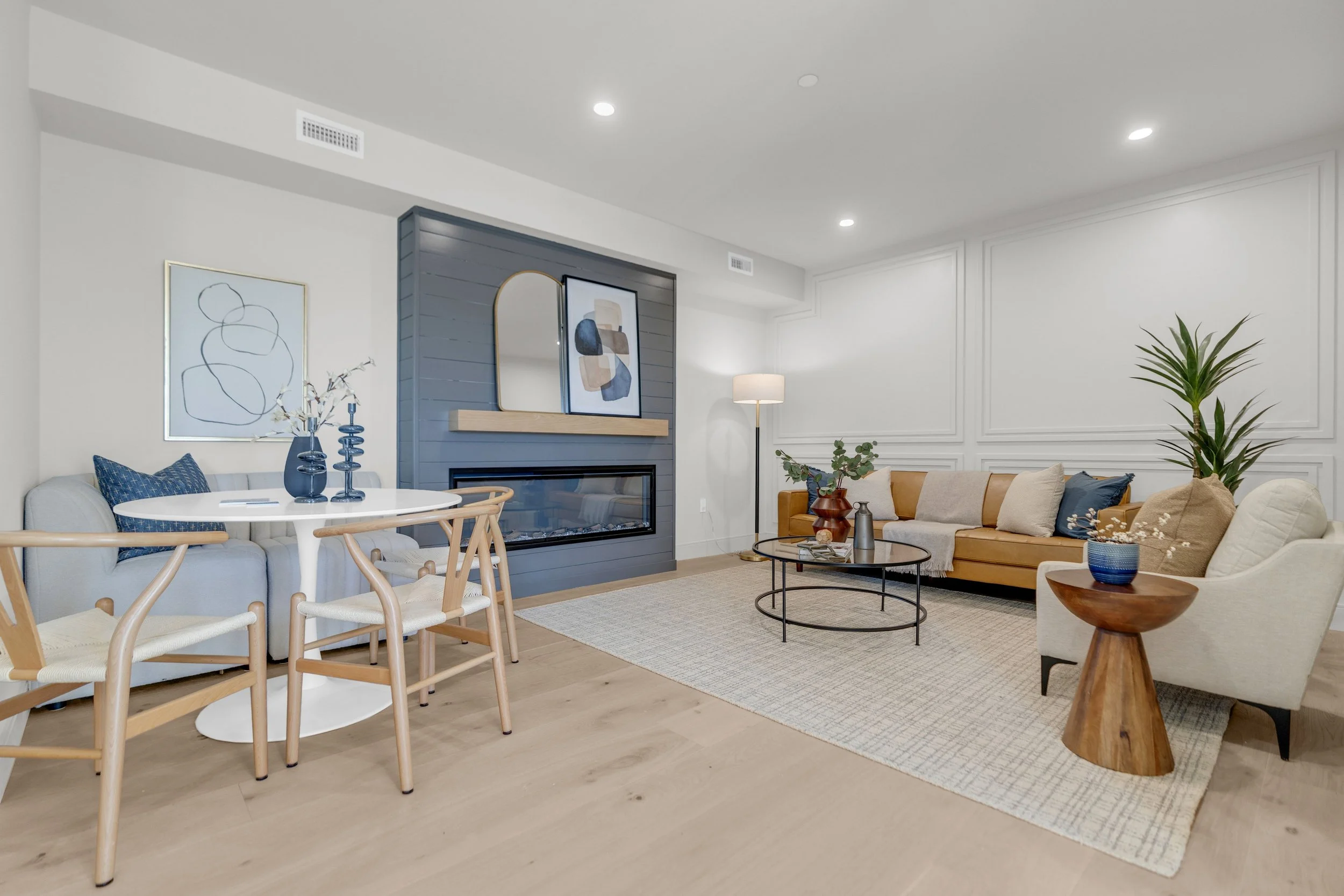 Living room with beige couch, white armchair, wood side table, black coffee table on light-colored rug, blue accents, and a fireplace with a decorative mirror and artwork above.