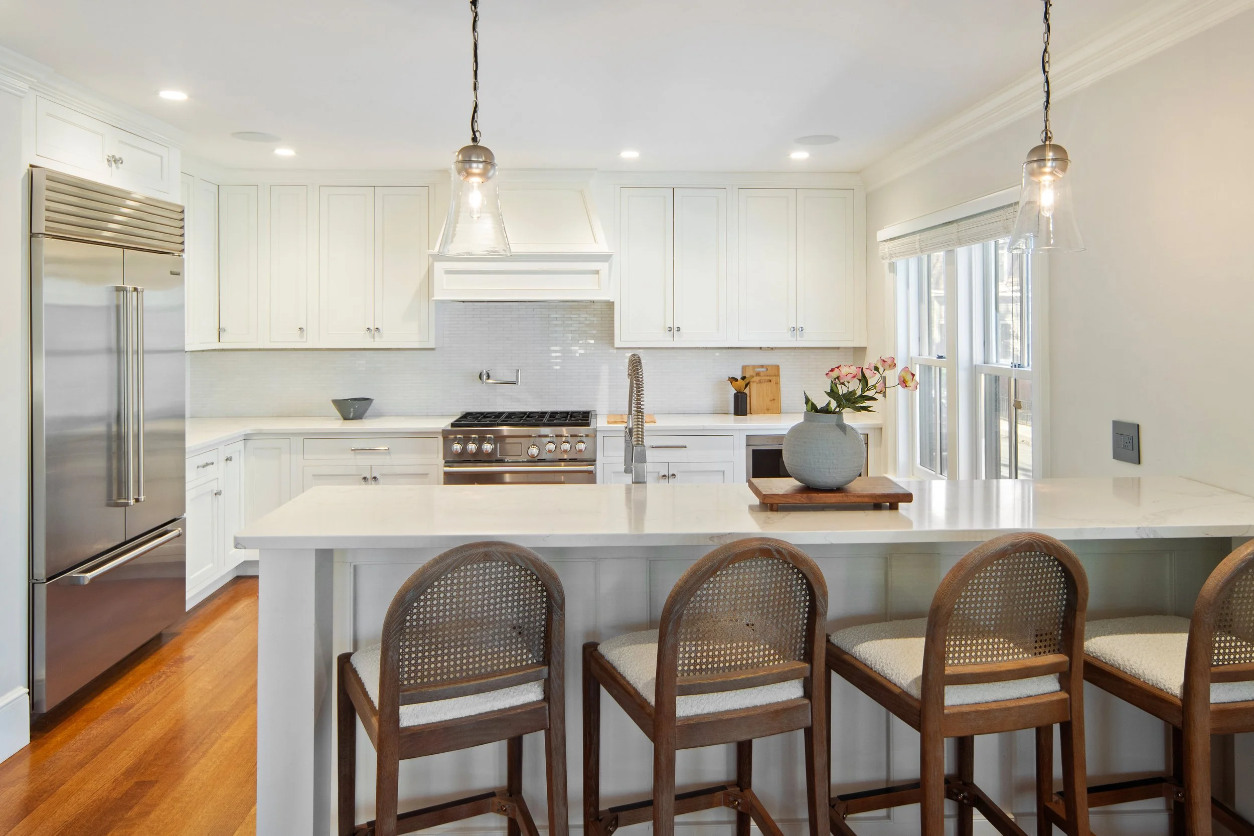 Modern kitchen with white cabinets, stainless steel refrigerator and range, marble island, pendant lights, and a flower arrangement on the counter.
