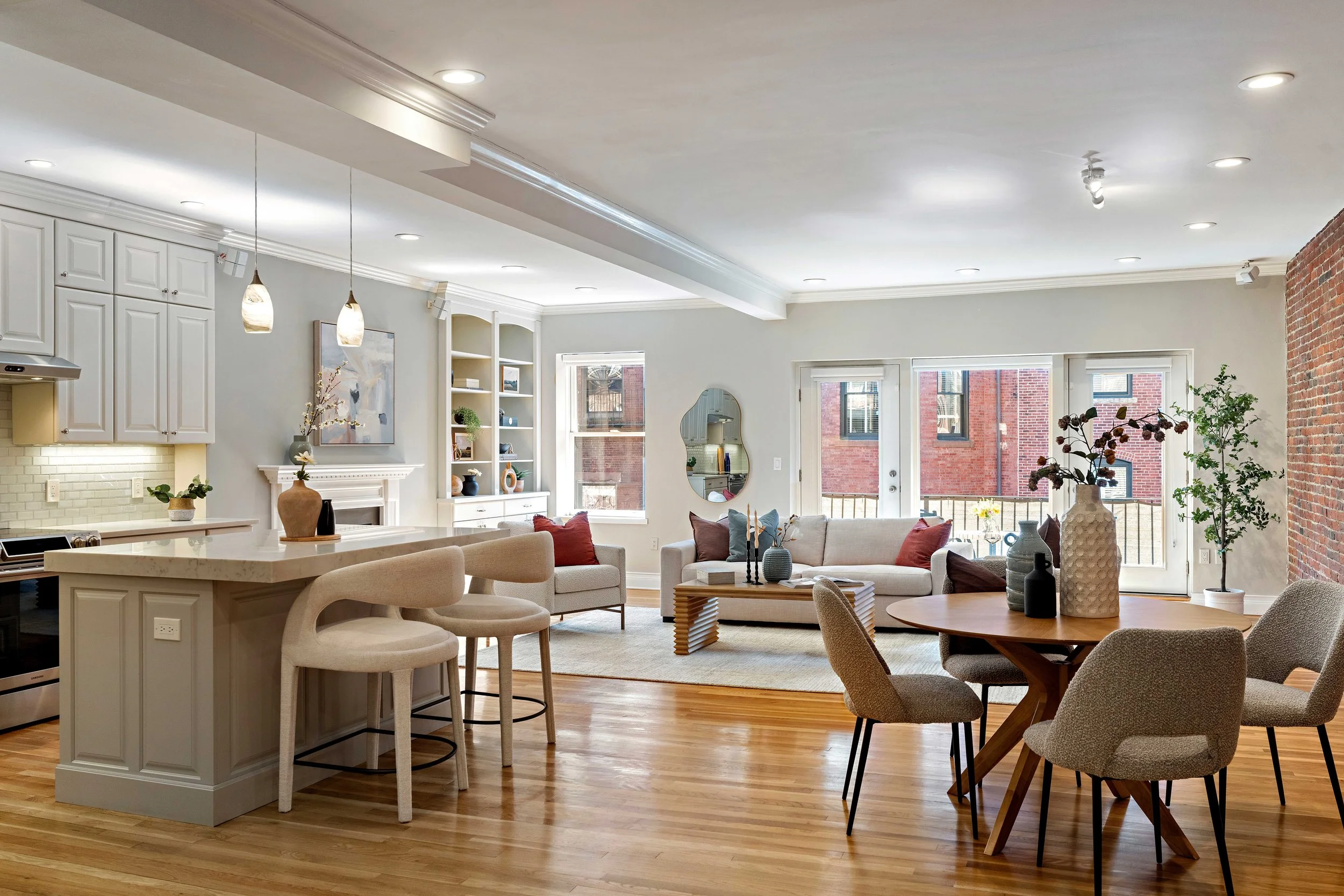Open-concept living room and kitchen with large windows, brick wall accent, modern furniture, plants, and pendant lighting.
