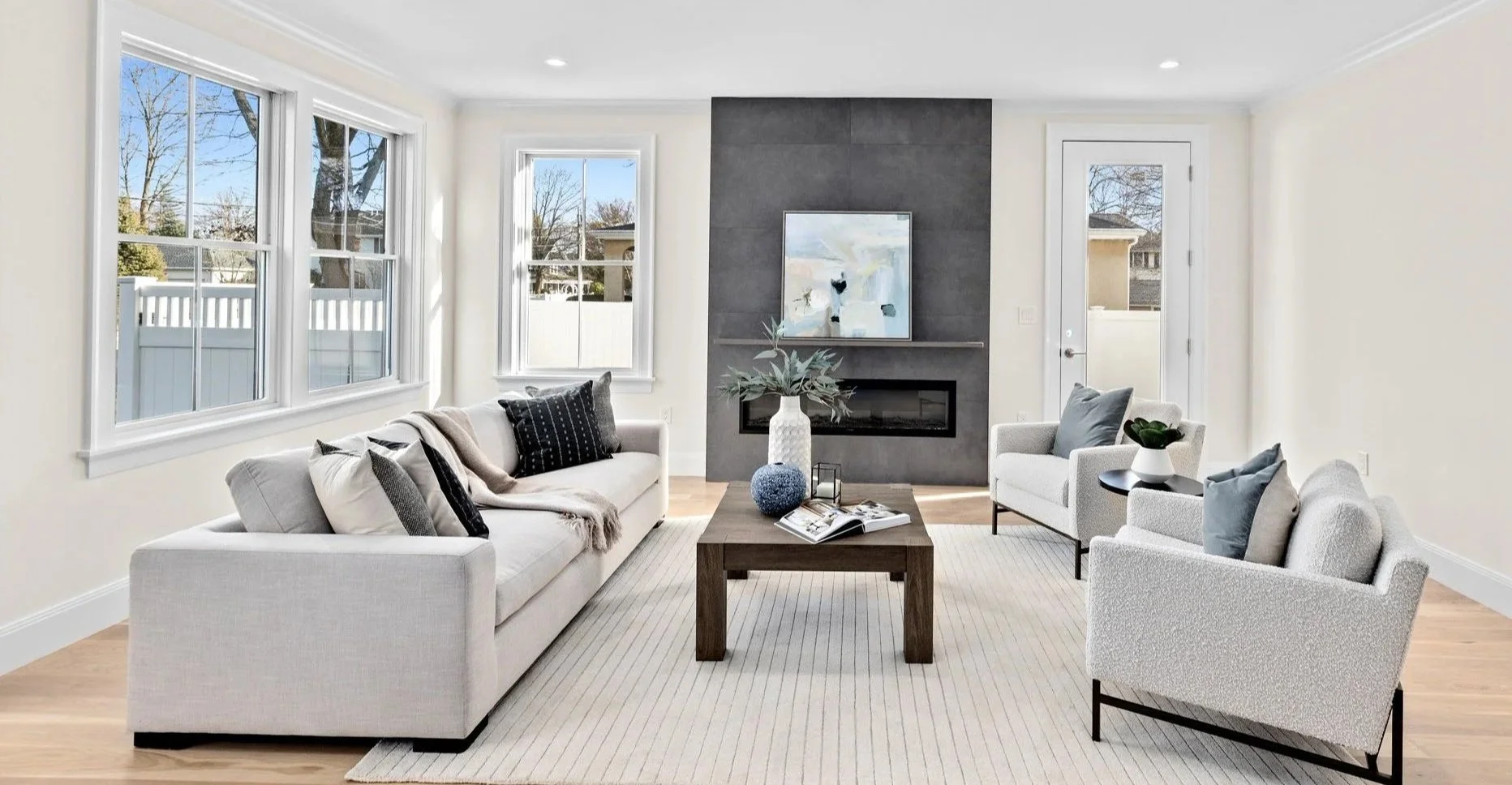Bright living room with white walls, three large windows, a fireplace with a dark gray surround, and seating including a white sofa and two white armchairs. There is a wooden coffee table with decorative objects and a painting above the fireplace, on a light-colored rug.