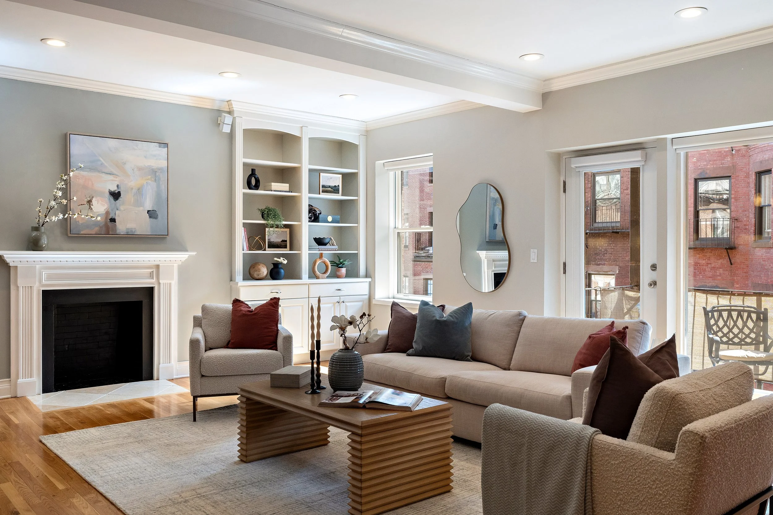 Bright living room with white walls, large windows, a fireplace, and beige furniture including a sofa and armchair, decorated with pillows and vases, with wooden flooring and a stacked wooden coffee table.
