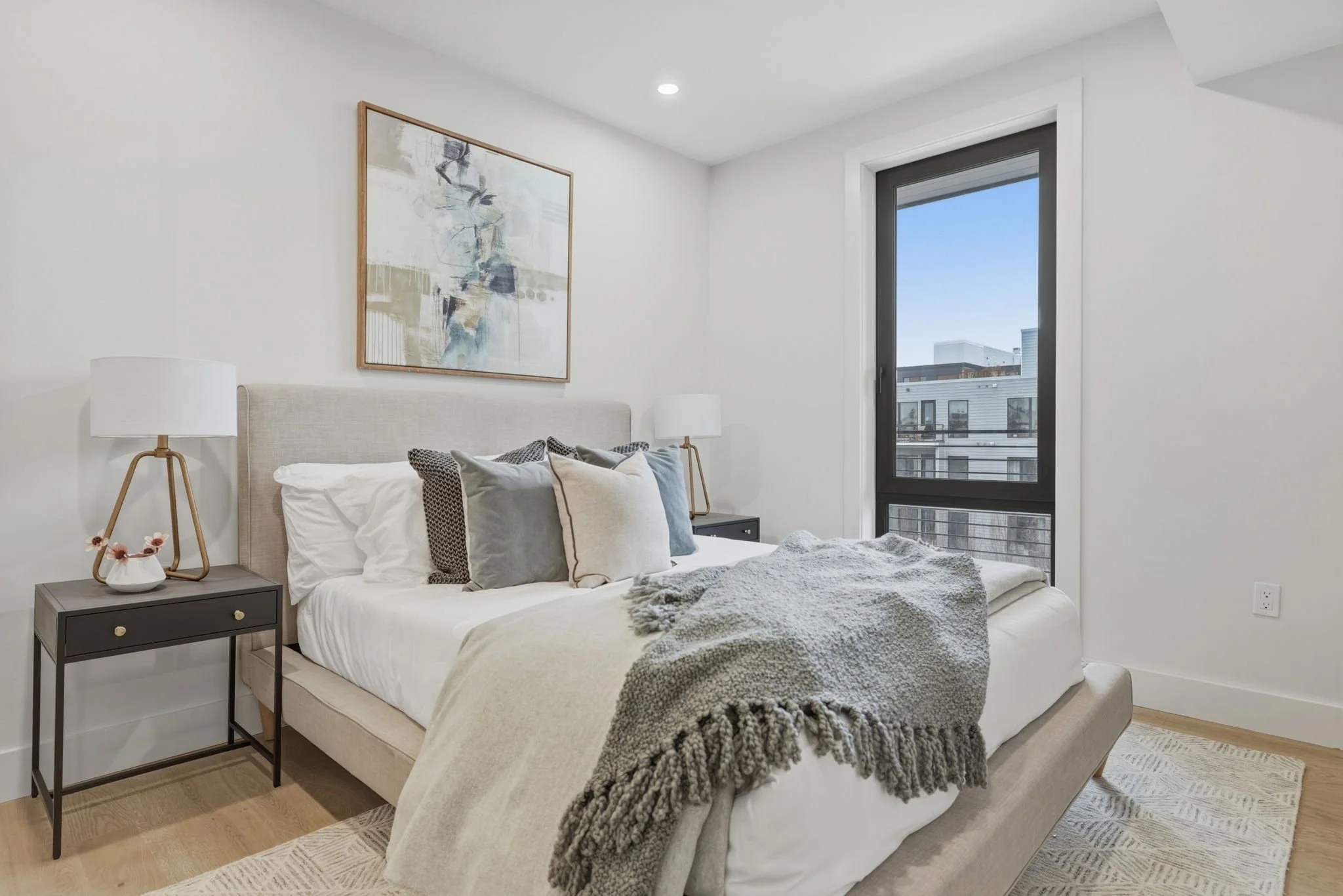 A modern bedroom with a bed, pillows, and a blanket, nightstand with a lamp, abstract artwork on the wall, large window showing city buildings, light-colored wooden floor, and light-colored walls.