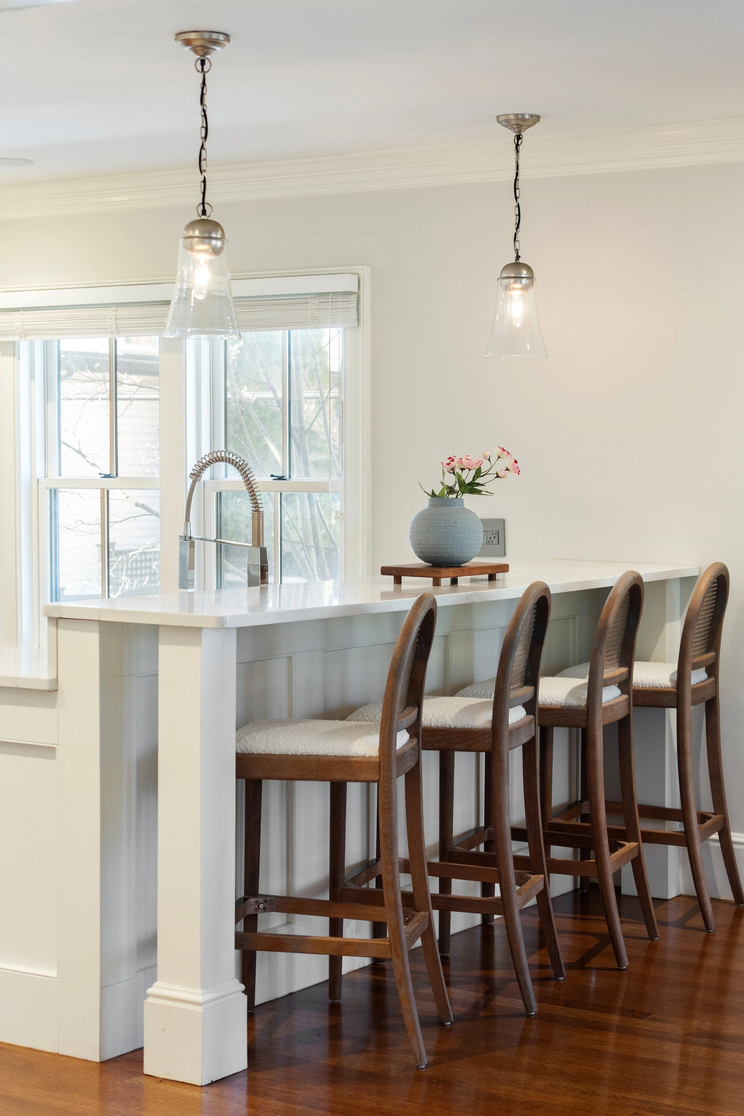 Kitchen island with four wooden chairs, a vase with pink flowers, windows with blinds, and pendant lights overhead.