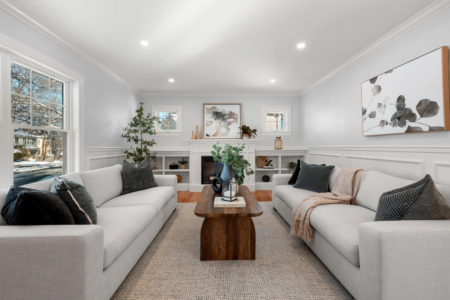 Living room with two white sofas, a wooden coffee table, indoor plants, decorative items, artwork, built-in shelves, and large window with a snowy outdoor scene.