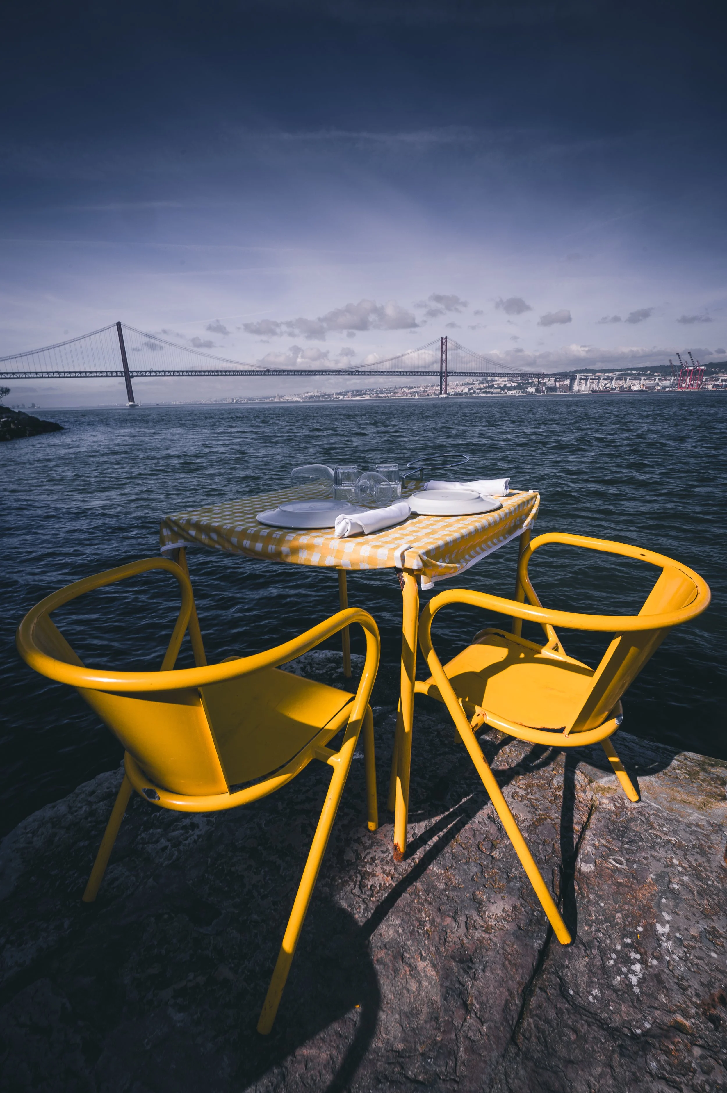 Ein Tisch mit gelben Stühlen und einer gelb-weiß karierter Tischdecke am Wasser, mit Blick auf eine Brücke und eine Stadt im Hintergrund.