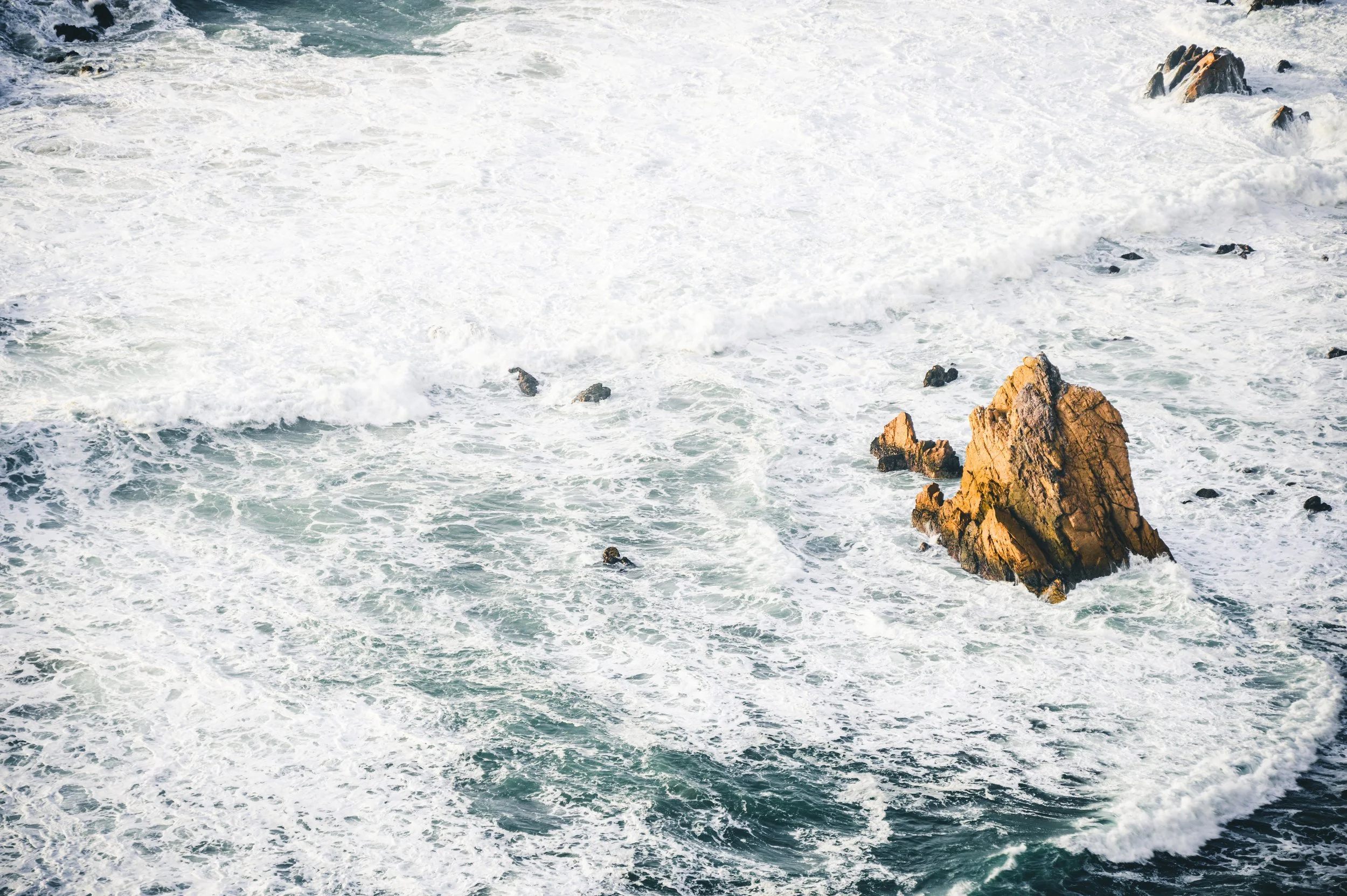 Das Bild zeigt eine Meeresküste mit großen Felsen im Wasser, Wellen schlagen gegen die Felsen und erzeugen Spritzwasser.