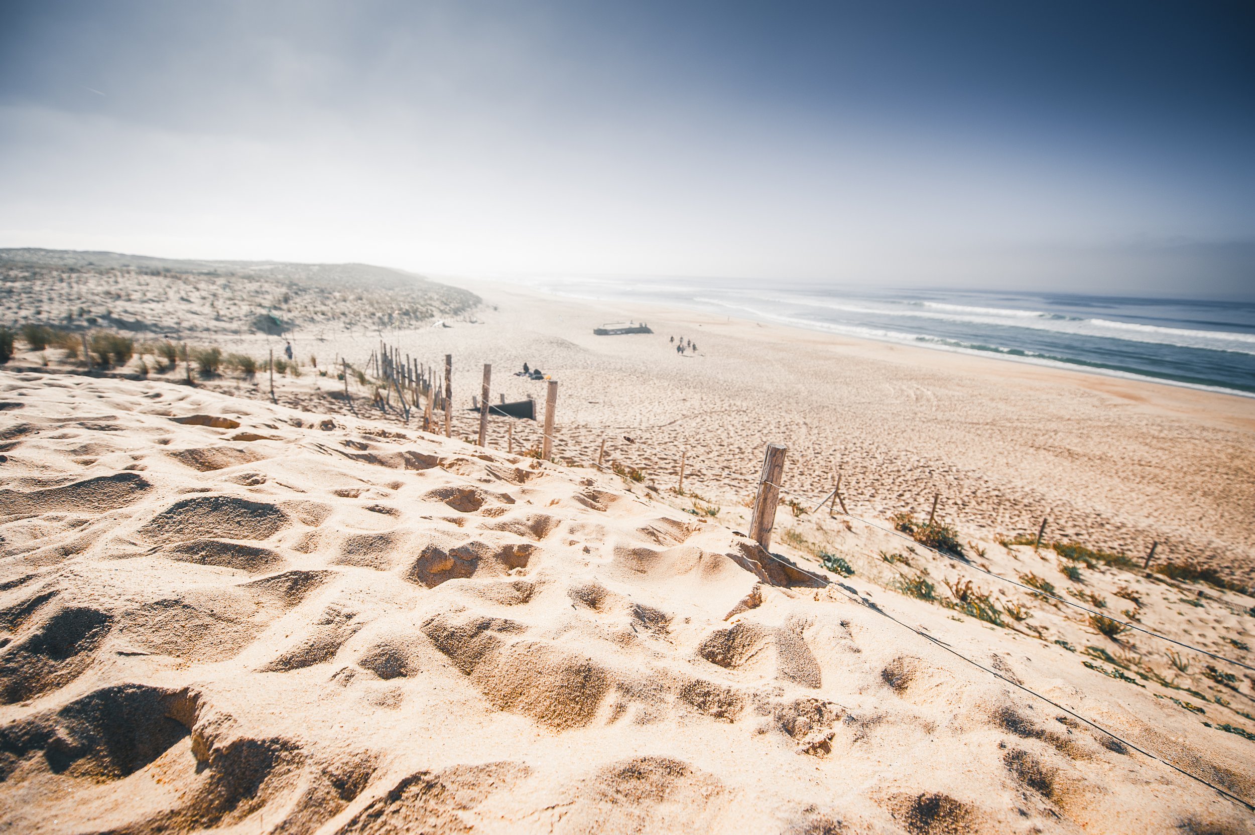 Weitwinkelaufnahme eines Strandes mit Sanddünen, einigen wenigen Menschen, Wellen und blauen Himmel.