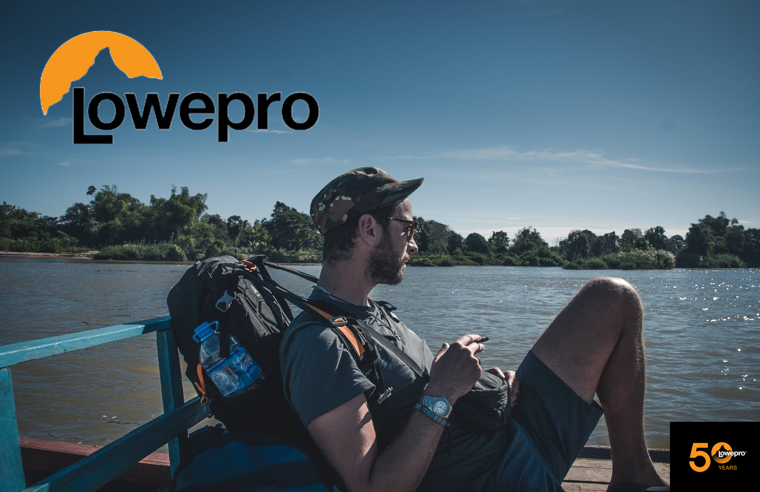 Ein Mann sitzt entspannt an einem Fluss mit bewusstem Blick auf den Wasser. Er trägt eine Mütze, eine Brille, eine Armbanduhr, einen Rucksack und ein T-Shirt. Der Himmel ist klar, und im Hintergrund sind Bäume sichtbar. Das Bild enthält das Lowepro-L