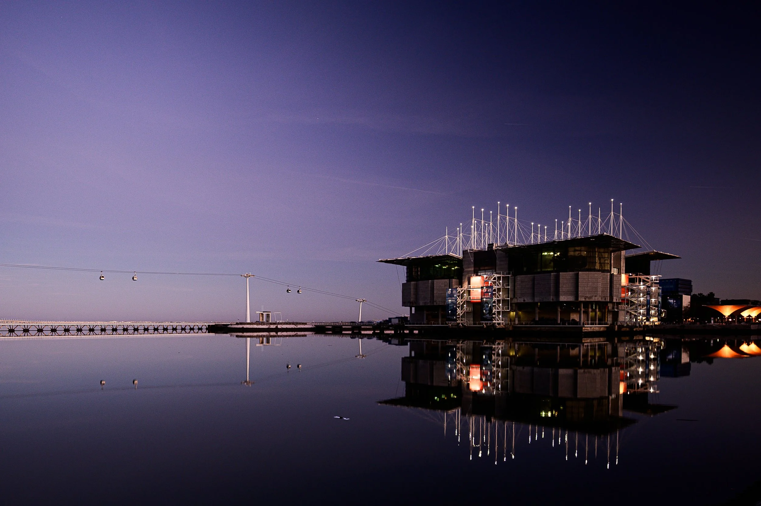 Moderne Gebäude am Wasser bei Nacht mit reflektiertem Himmel und einem Kabelbahn auf der linken Seite.