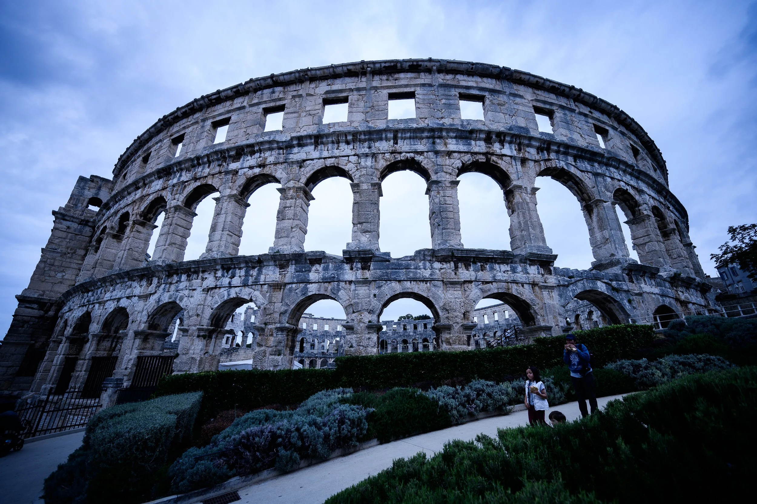 Das Bild zeigt das römische Amphitheater in Verona bei bewölktem Himmel, mit zwei Personen im Vordergrund, einer Frau und einem Kind, die auf einem Weg stehen.