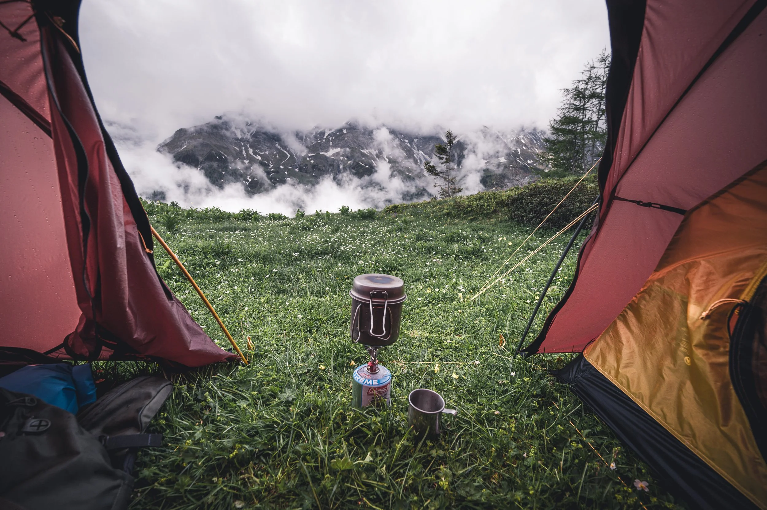 Blick aus einem Zelt in die Natur, mit grüner Wiese, Bergen und Wolken im Hintergrund, Campingausrüstung im Vordergrund.
