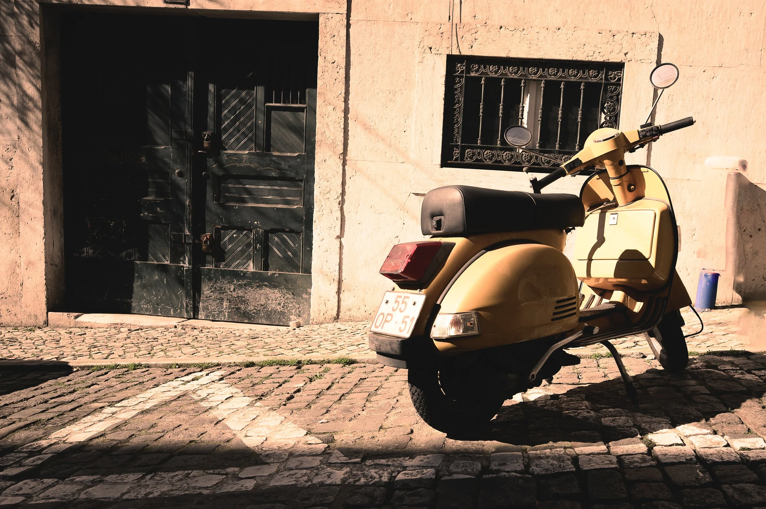 Ein gelber Motorroller steht auf einem gepflasterten Bürgersteig vor einer alten, schwarzen, Holztür und einem Fenster mit schmiedeeisernem Gitter. Die Szene ist sonnig, mit Schatten auf dem Boden.