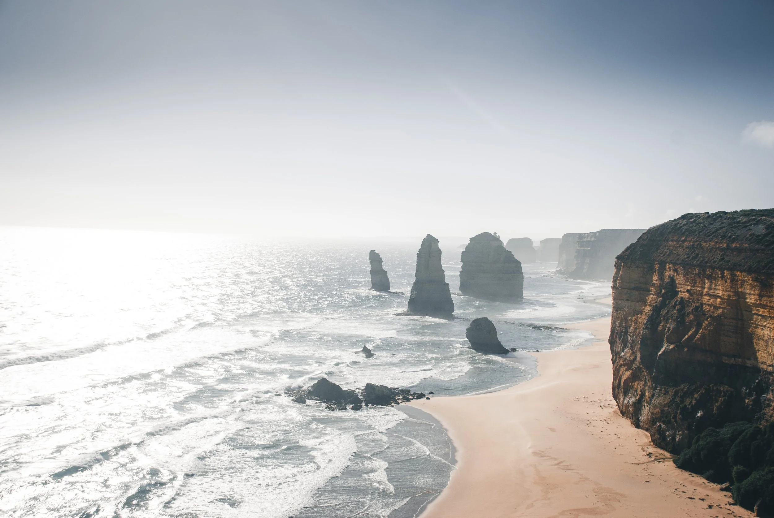 Eine Küstenlandschaft mit Felsen im Meer, Sandstrand und Klippen bei Sonnenlicht.