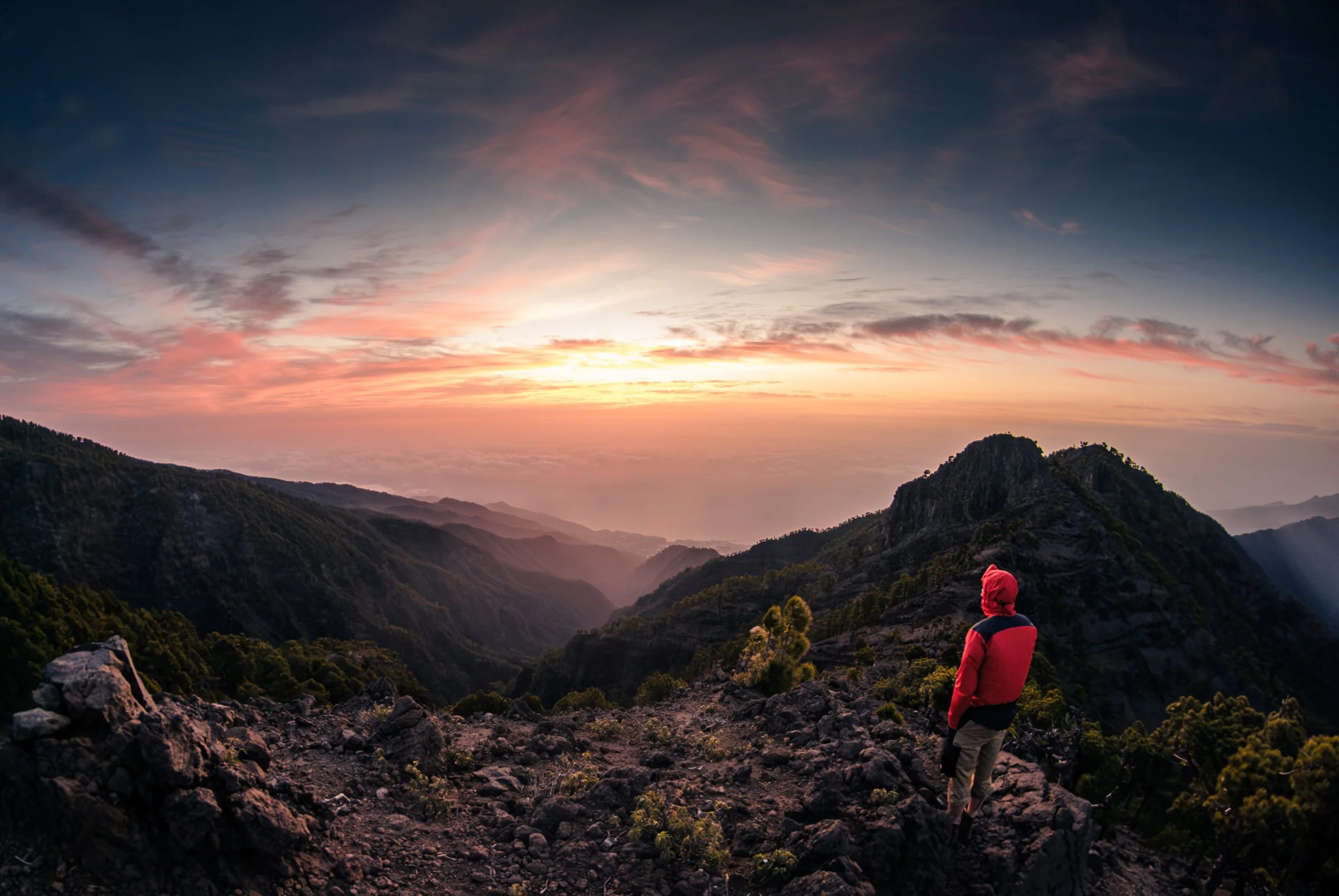 Ein Wanderer in einer roten Jacke und Mütze steht auf felsigem Terrain und blickt auf ein Tal mit Bergen bei Sonnenuntergang, im Himmel sind Wolken in warmen Farben.