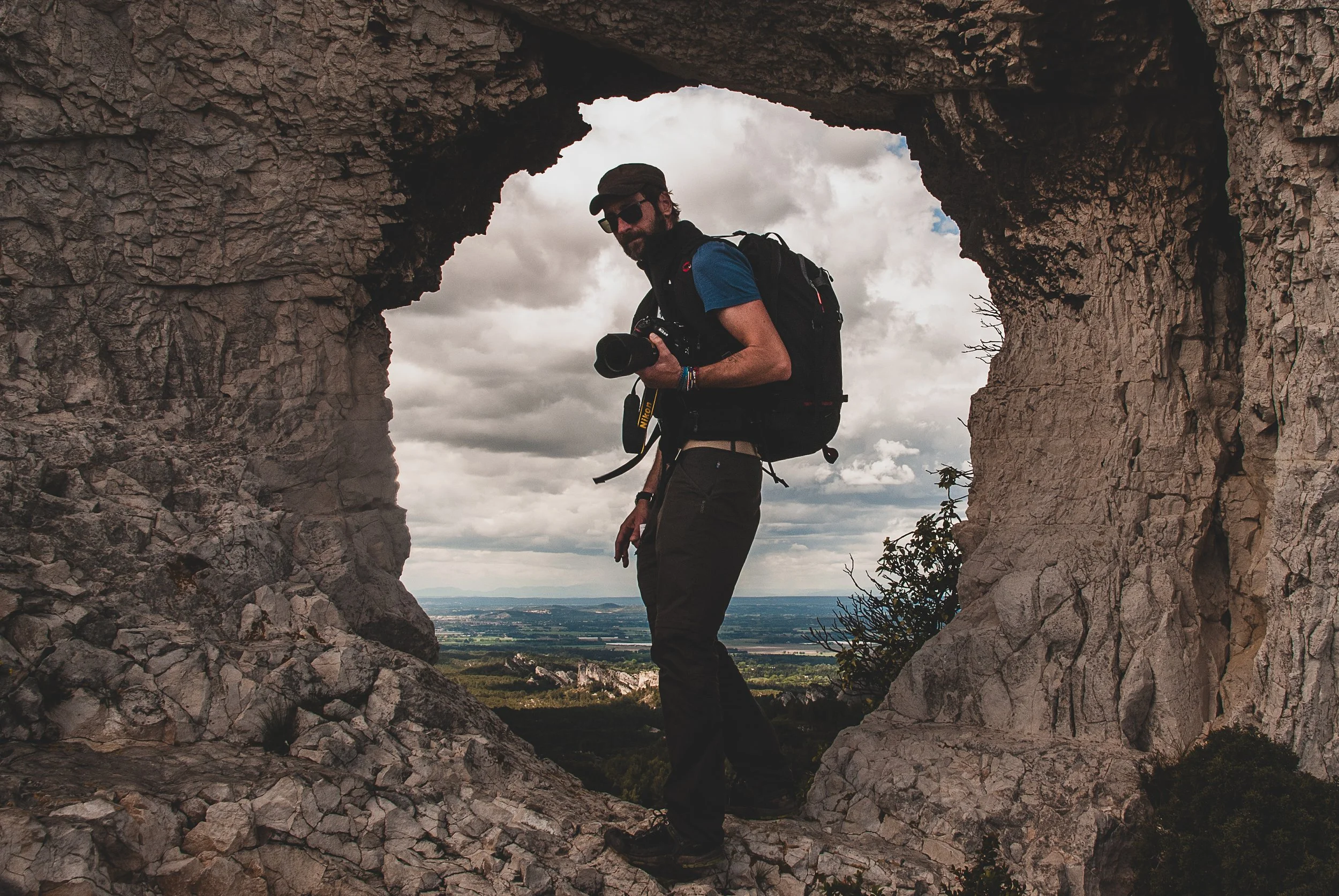 Ein Mann mit Kamera steht in einer Felsöffnung auf einer Berglandschaft, trägt Rucksack und Sonnenbrille, bei bewölktem Himmel.