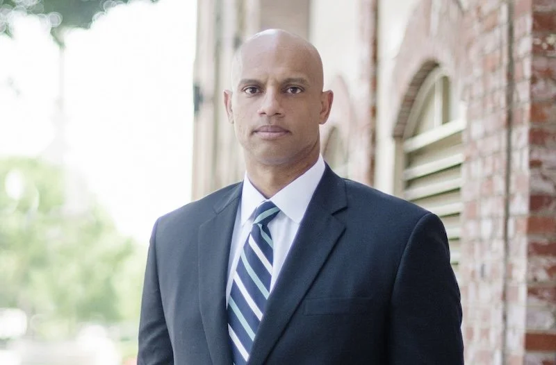 A bald man in a dark suit and striped tie standing outdoors in front of a brick building with green trees in the background.