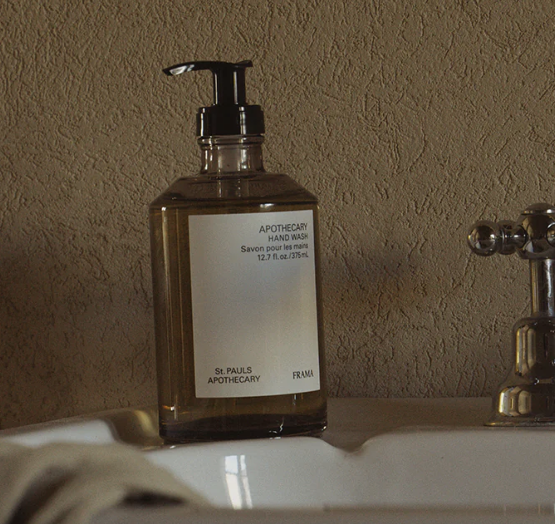 A clear plastic bottle of hand wash soap with black pump, labeled 'APOTHECARY HAND WASH, savon pour les mains, 12.7 fl oz, 375mL, St. PAULS APOTHECARY, FRANCE', placed on a bathroom sink next to a chrome faucet with three round handles.