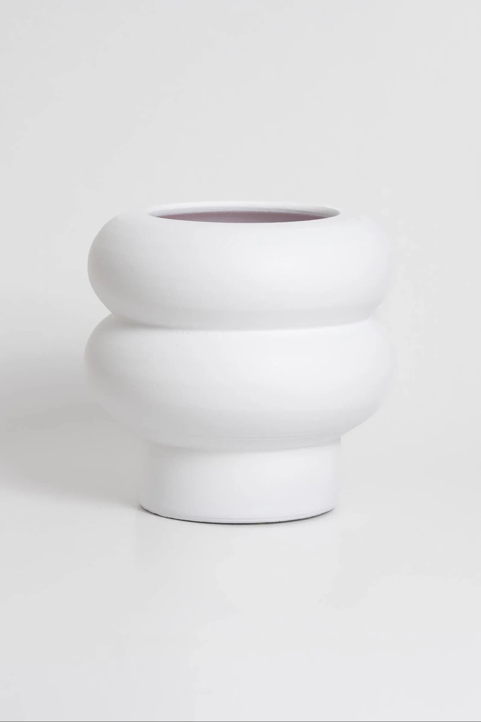 Minimalist white ceramic vase with a rounded, layered design on a white surface against a white background.