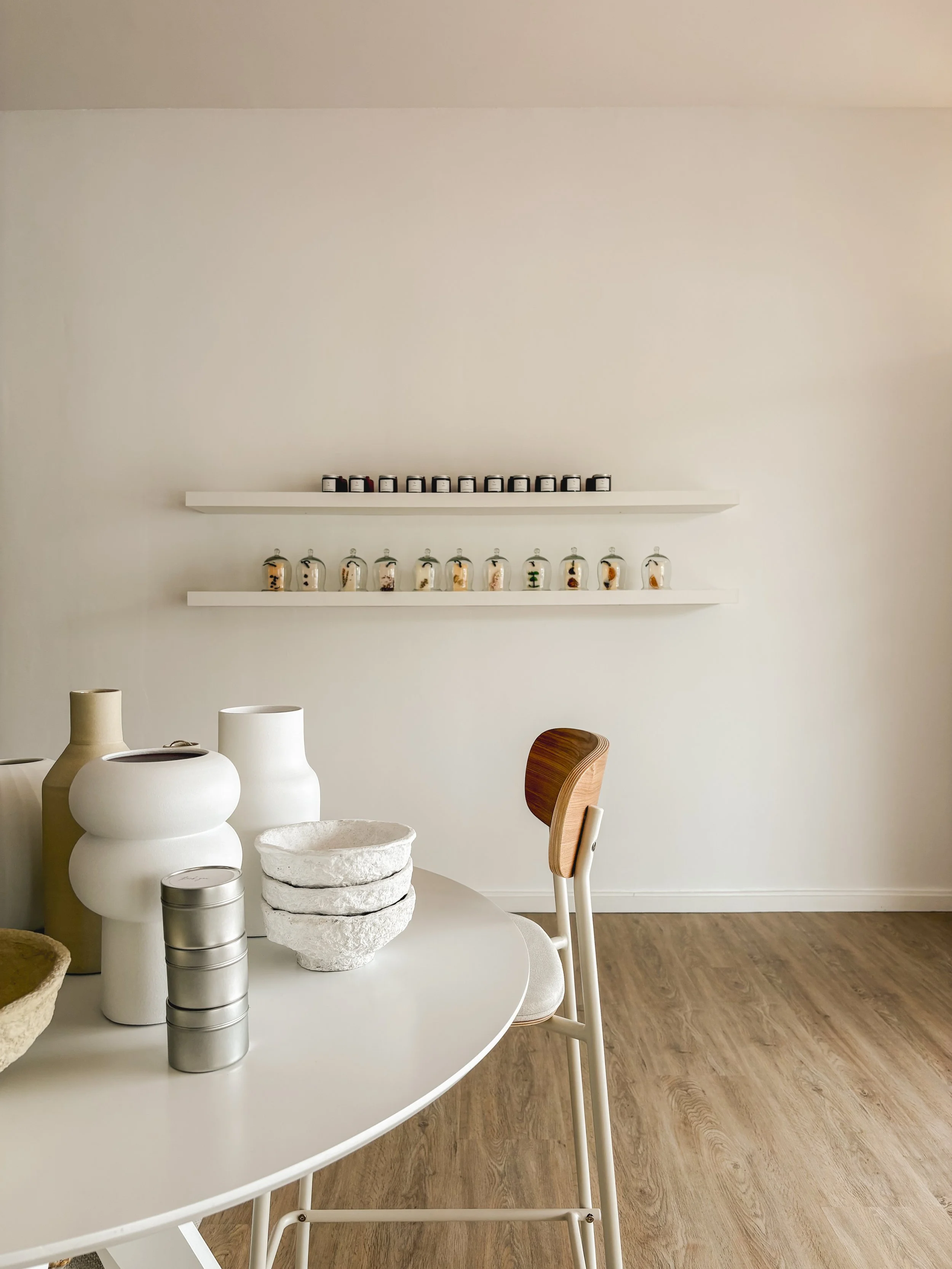 A minimalist room with white walls, wooden floor, and modern decor including tall vases, bowls, and a stacking container on a white round table. Two white shelves with jars and glass containers are mounted on the wall.