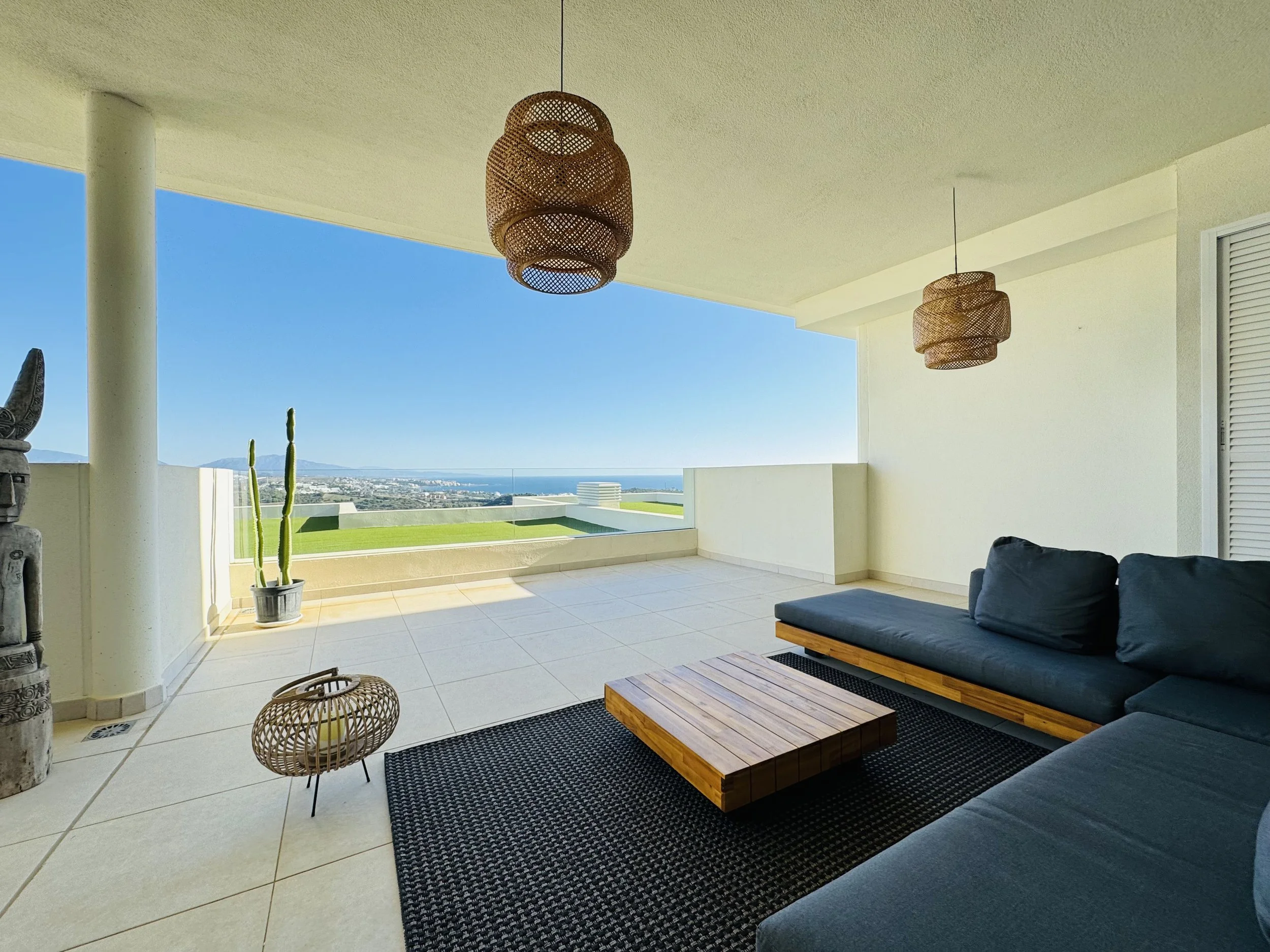 A spacious balcony with white tiled flooring, black cushioned outdoor sofa, wooden coffee table, hanging wicker lamps, a tall potted cactus, and an ocean view with a clear blue sky in the background.