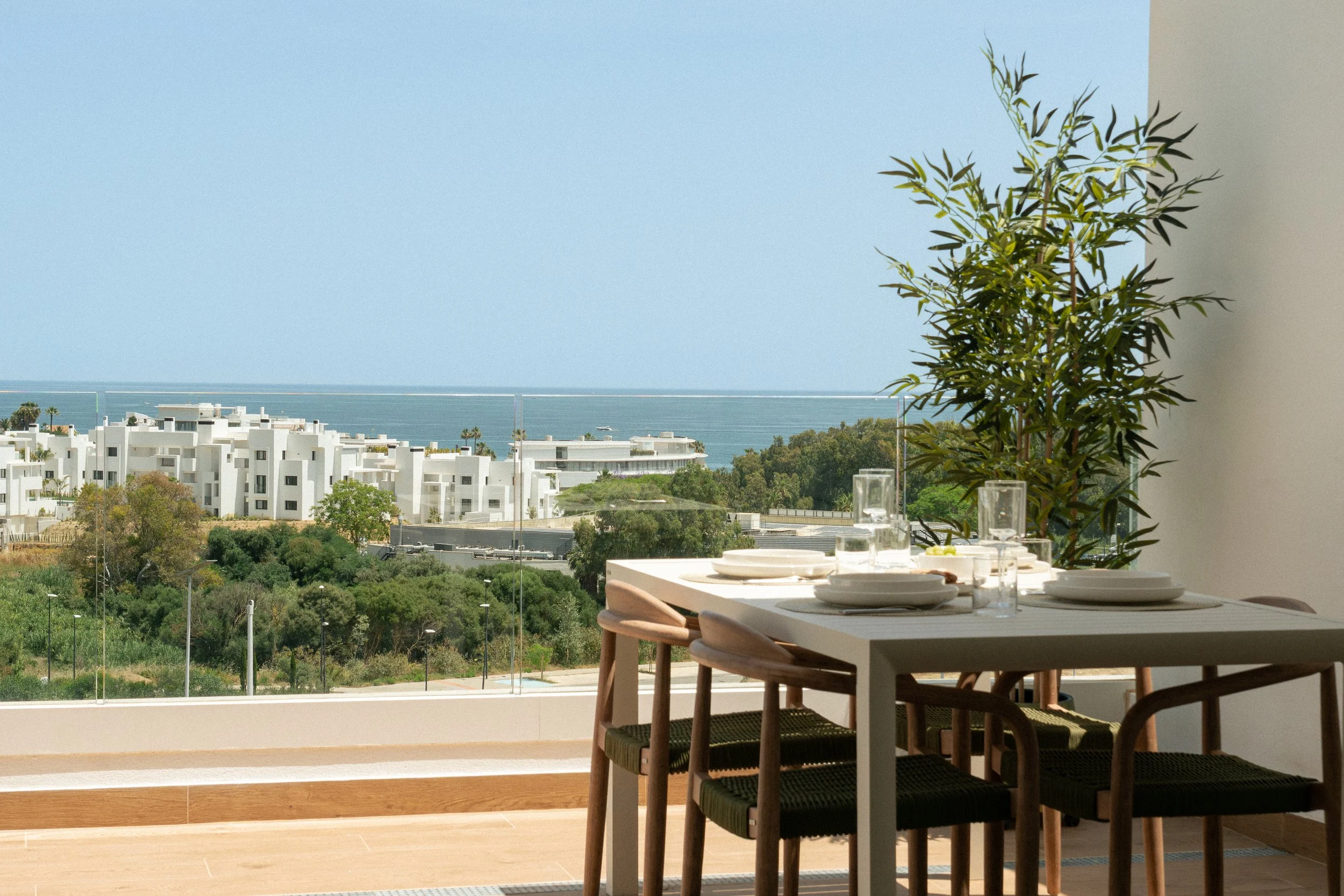Dining table set for a meal overlooking a scenic coastal view with white modern buildings, green trees, and the ocean in the background.