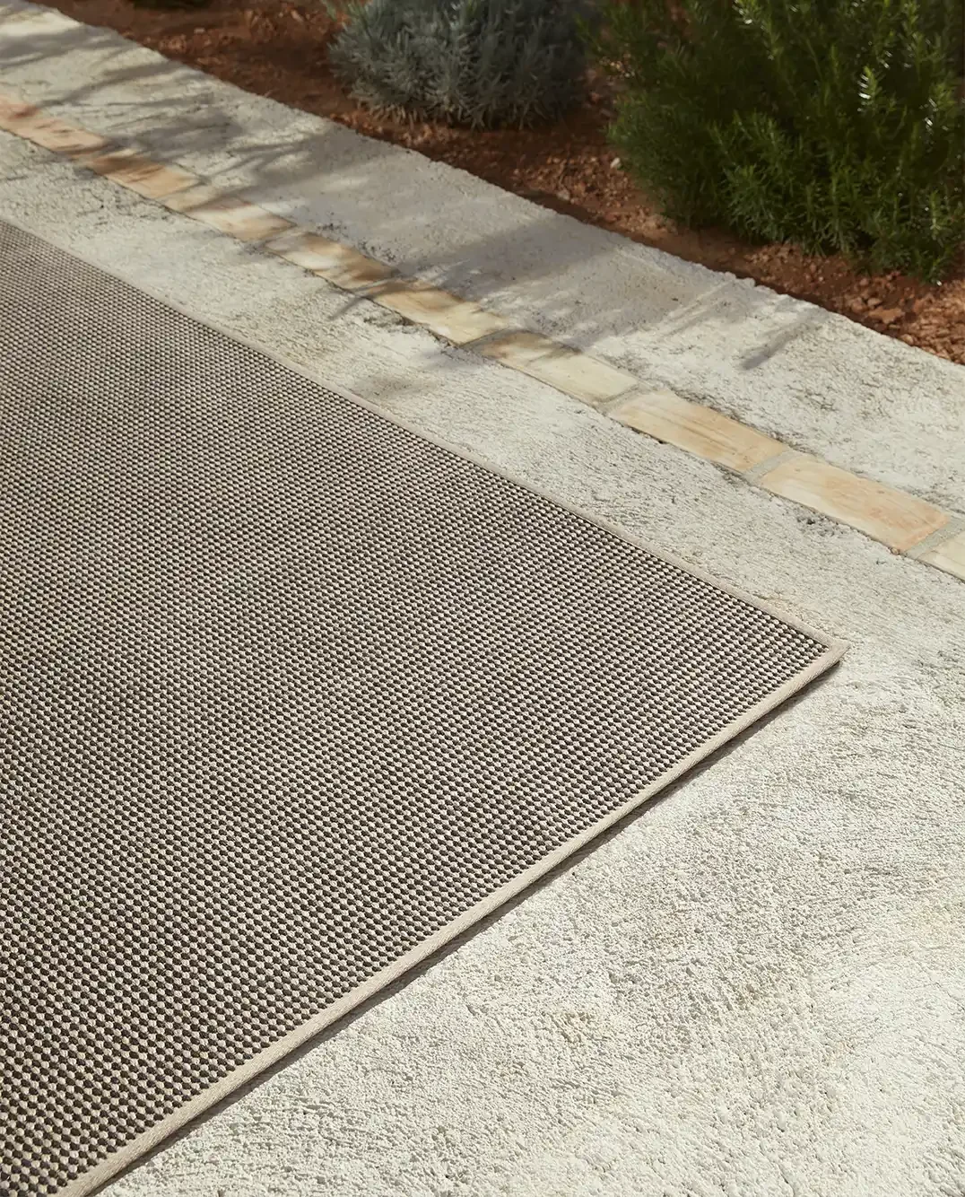 Outdoor concrete walkway with a patterned beige outdoor rug, bordered by a garden bed with shrubs and mulch.