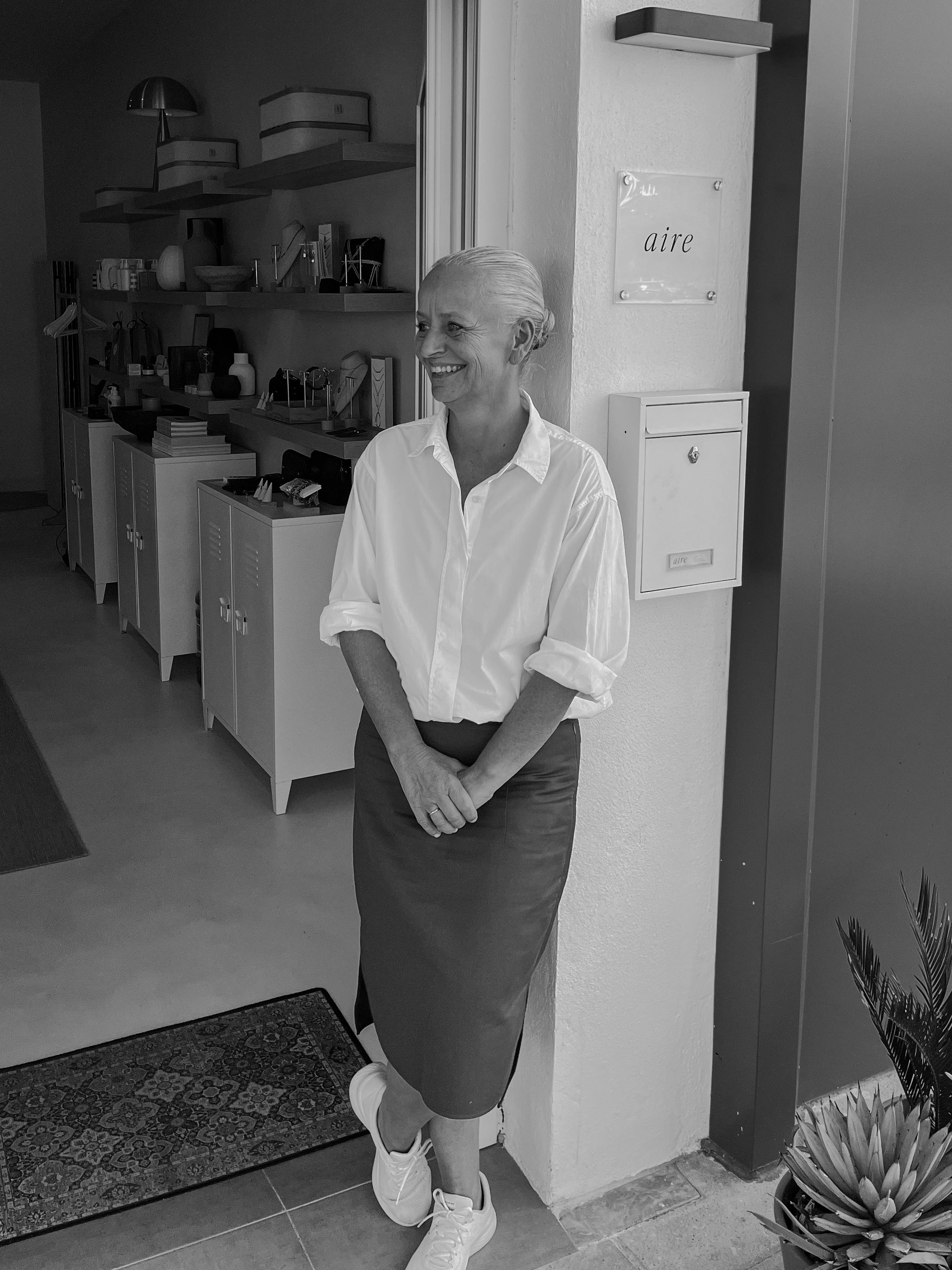 A woman standing with her back against a wall in a room, smiling, wearing a white shirt, a dark skirt, and sneakers.