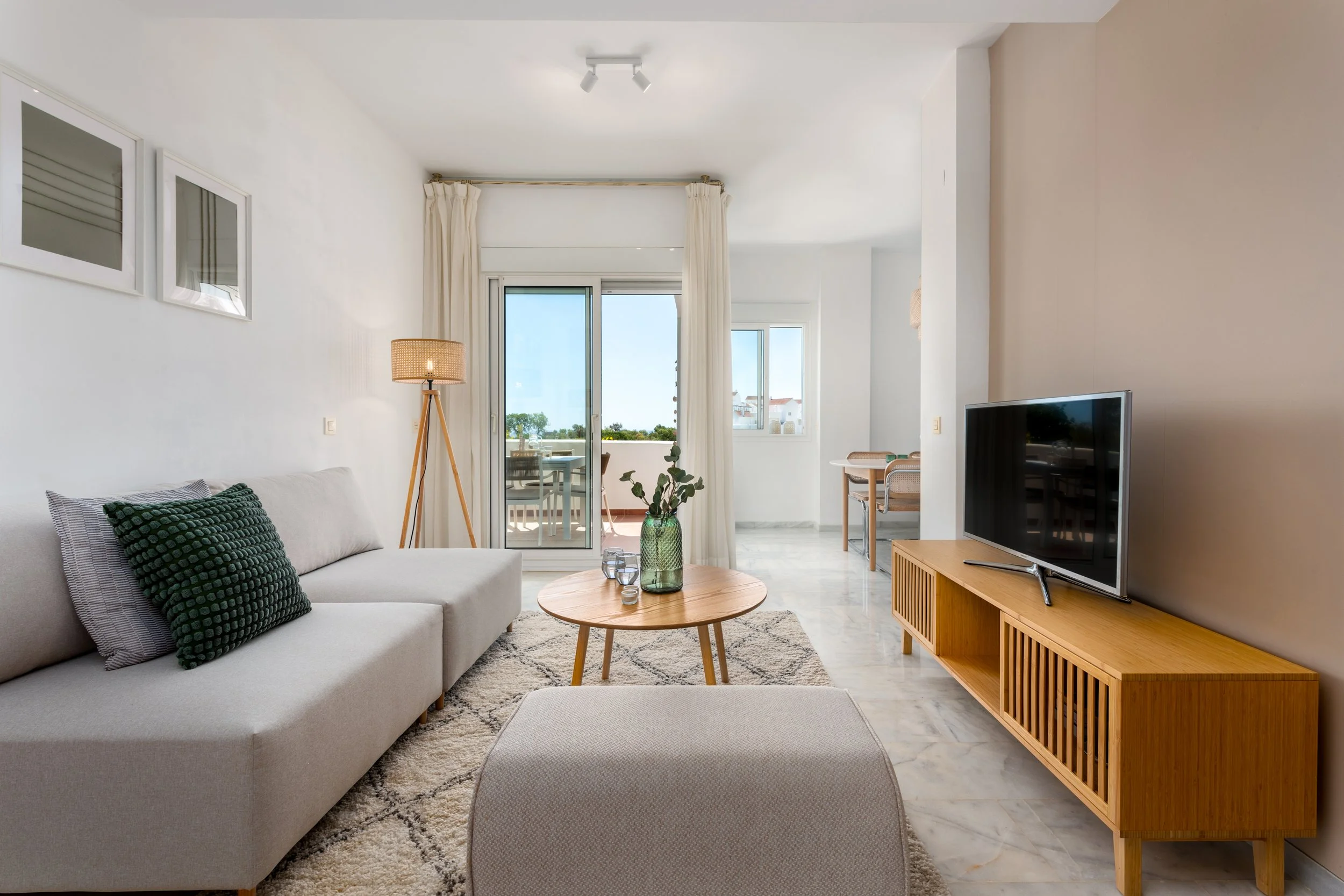 Living room with a sofa, flat-screen TV on a wooden stand, round coffee table with green vase, seating area on balcony with outdoor table and chairs, and cream curtains.
