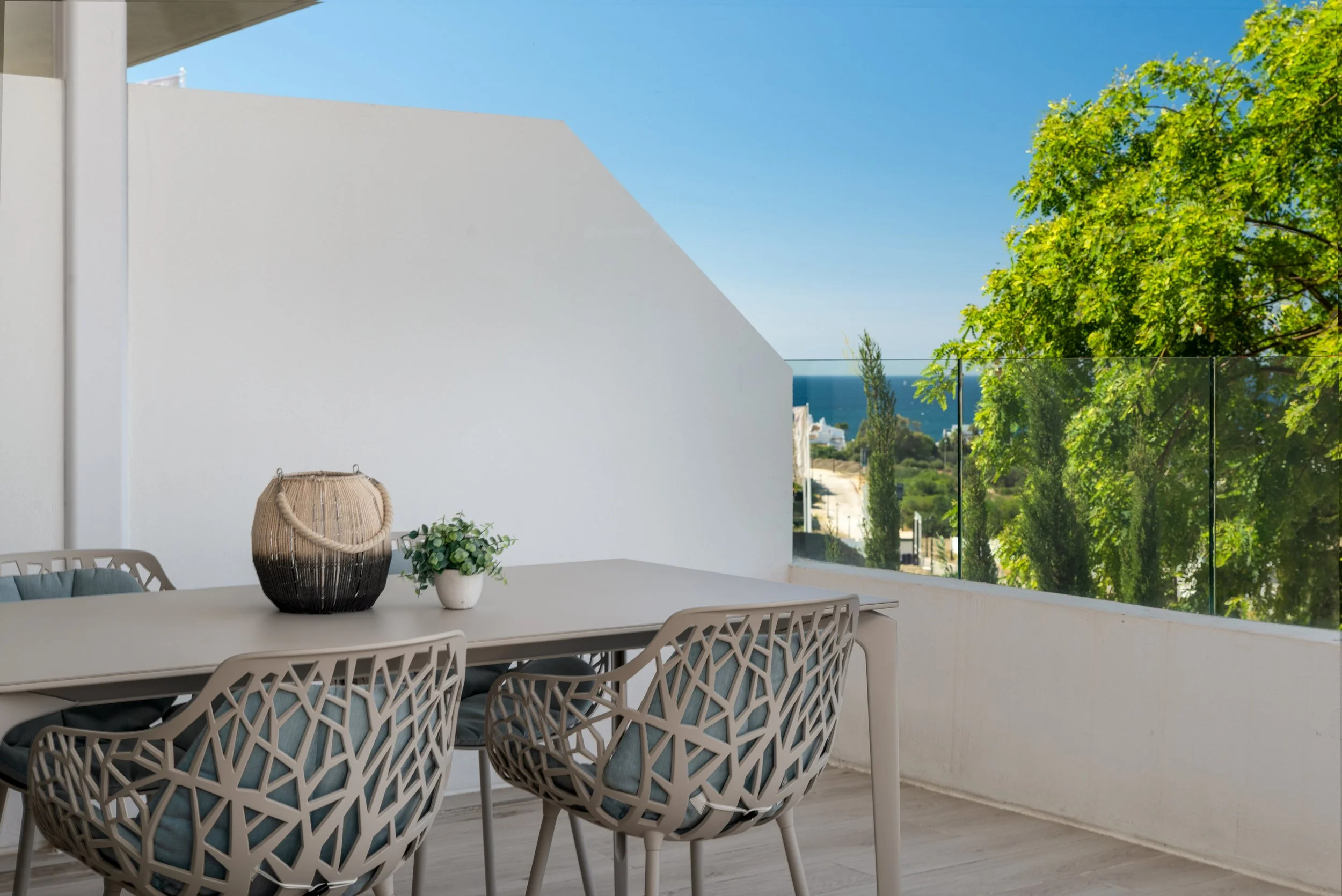 Outdoor balcony with a white dining table, four modern chairs with geometric backs, a basket, and a small plant. In the background, trees, a street, and the ocean are visible under a clear blue sky.