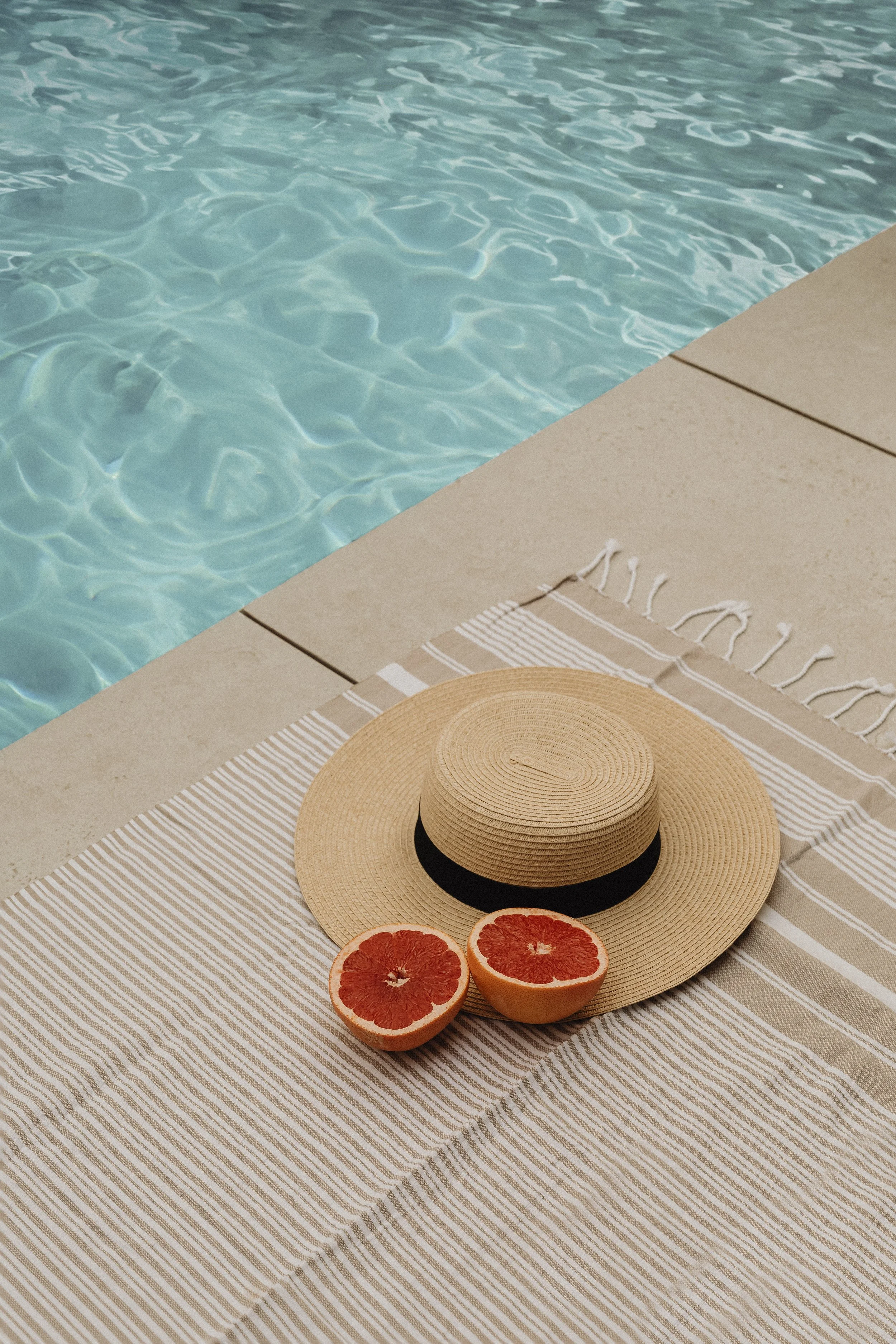 A straw hat with a black band, two halves of a grapefruit, placed on a striped beige and white towel by a swimming pool.