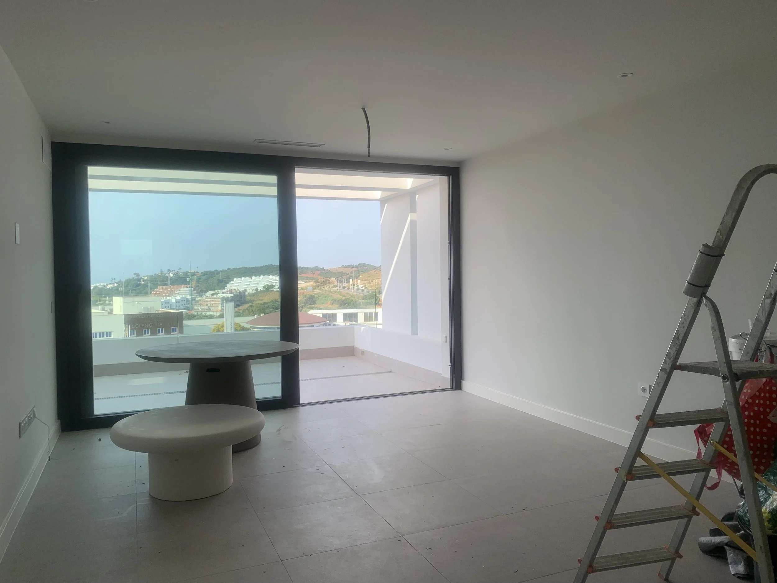 Empty room with sliding glass doors leading to a balcony, a small round table, a bench in the room, and a ladder on the side, under construction or renovation.