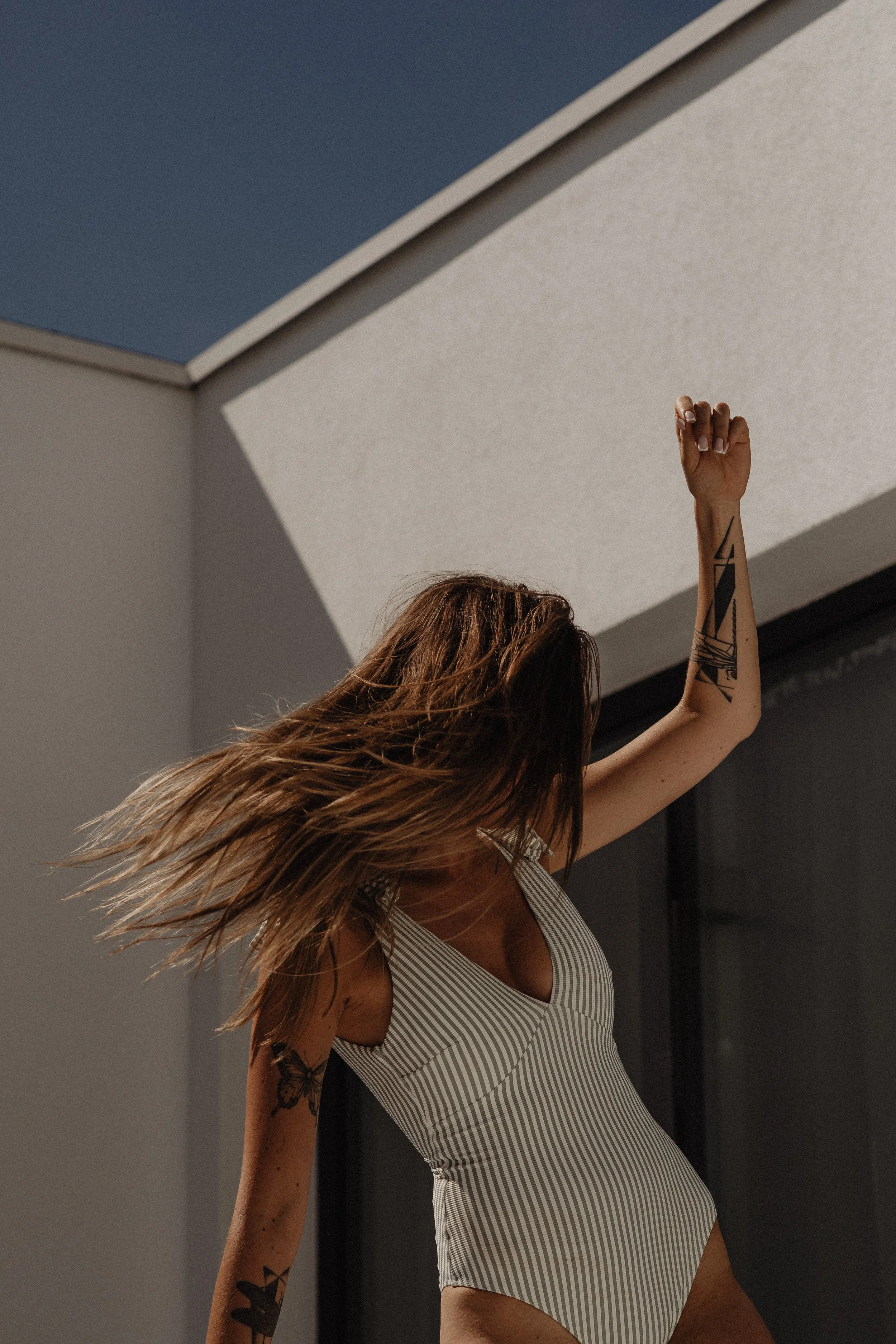 A woman with long hair, tattoos on her arm, wearing a striped swimsuit, is dancing or posing outdoors in sunlight with her hair moving.